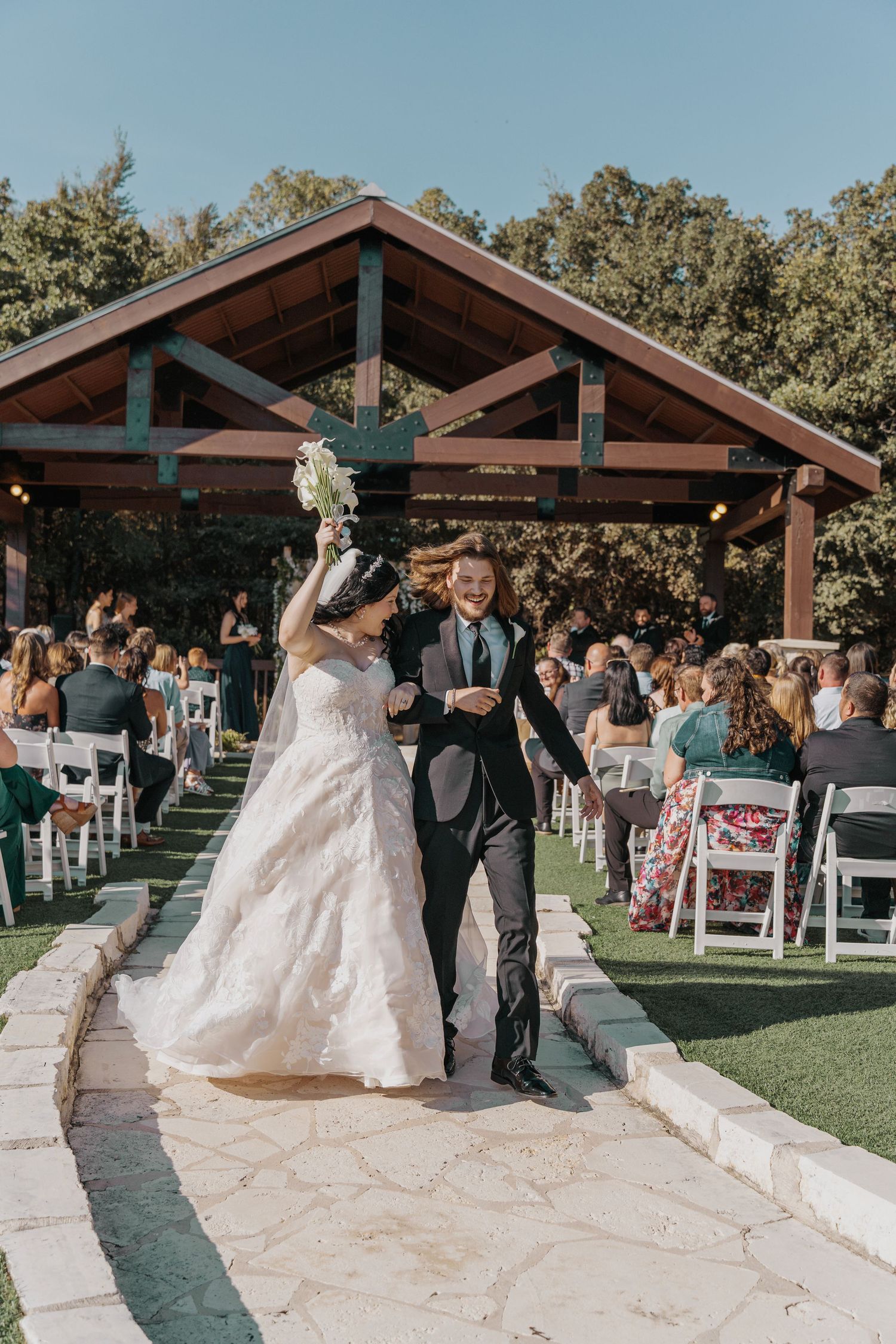 Joyful newlyweds walk down outdoor wedding aisle, bride in white gown holding bouquet aloft, groom in dark suit, guests seated.