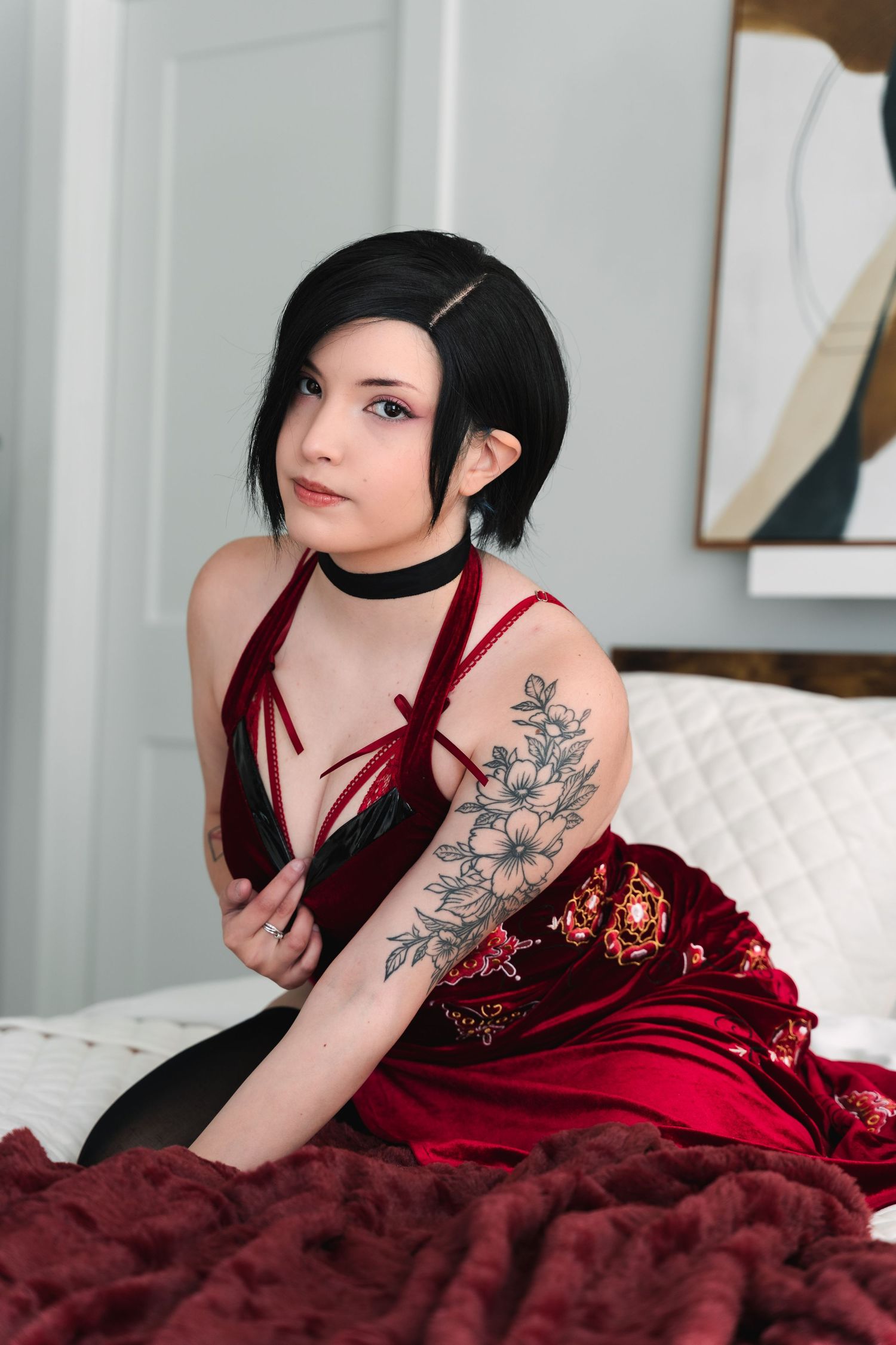 Model in red velvet dress with floral arm tattoo poses on white bedding.