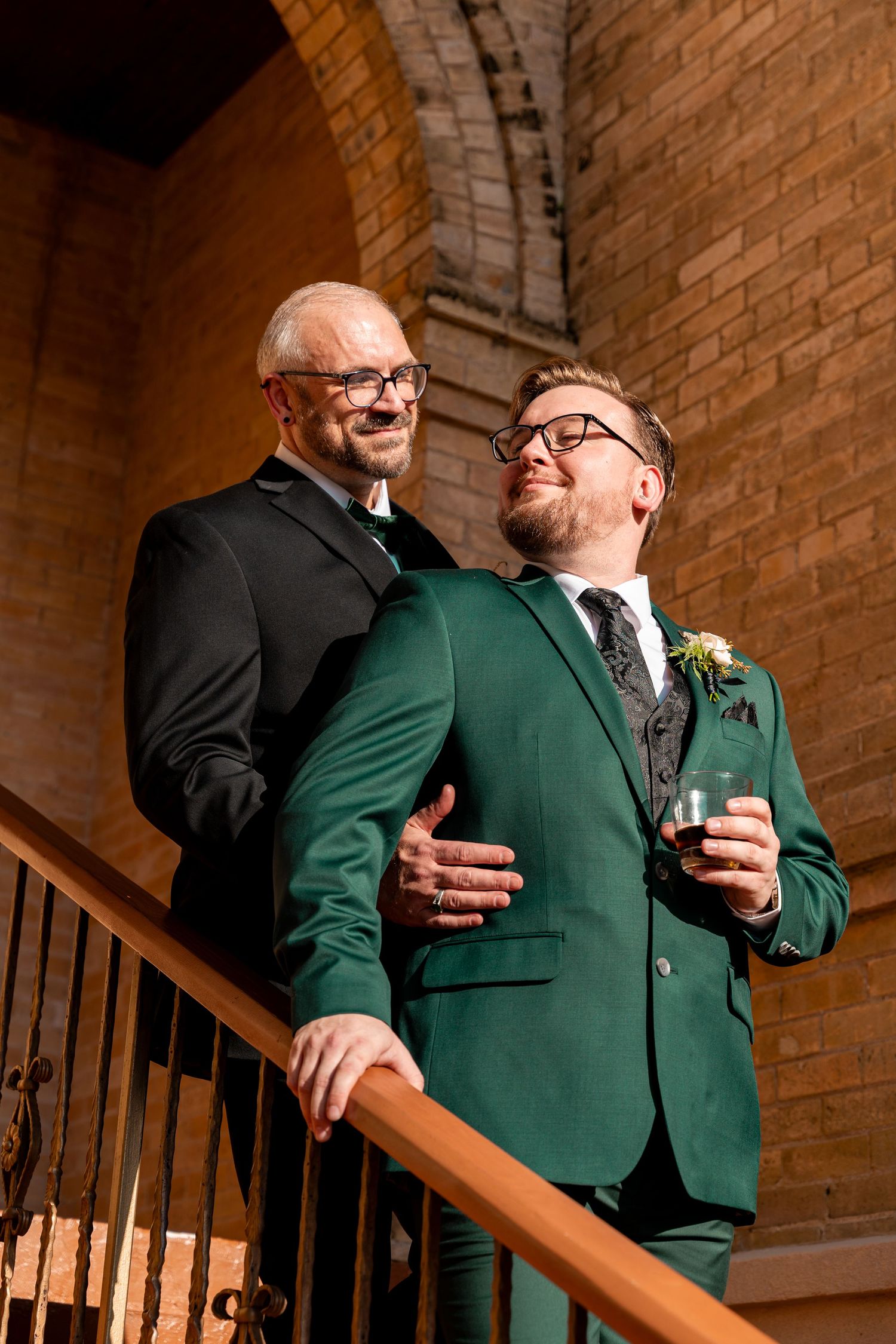 Two people in green and black formal wear share candid moments on wooden stairs.