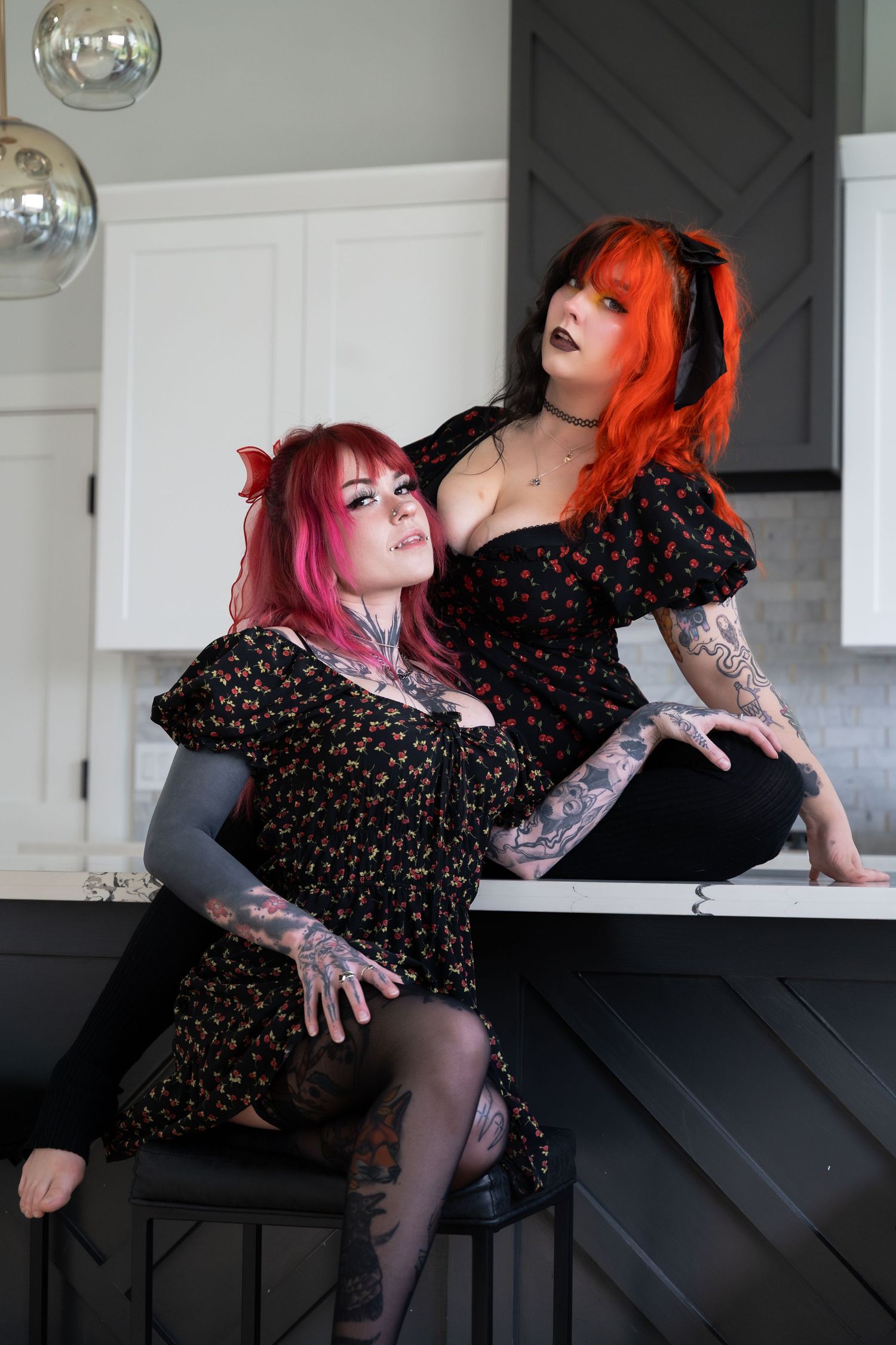 Two tattooed women with vibrant red and pink hair pose in floral black dresses in a modern kitchen setting.