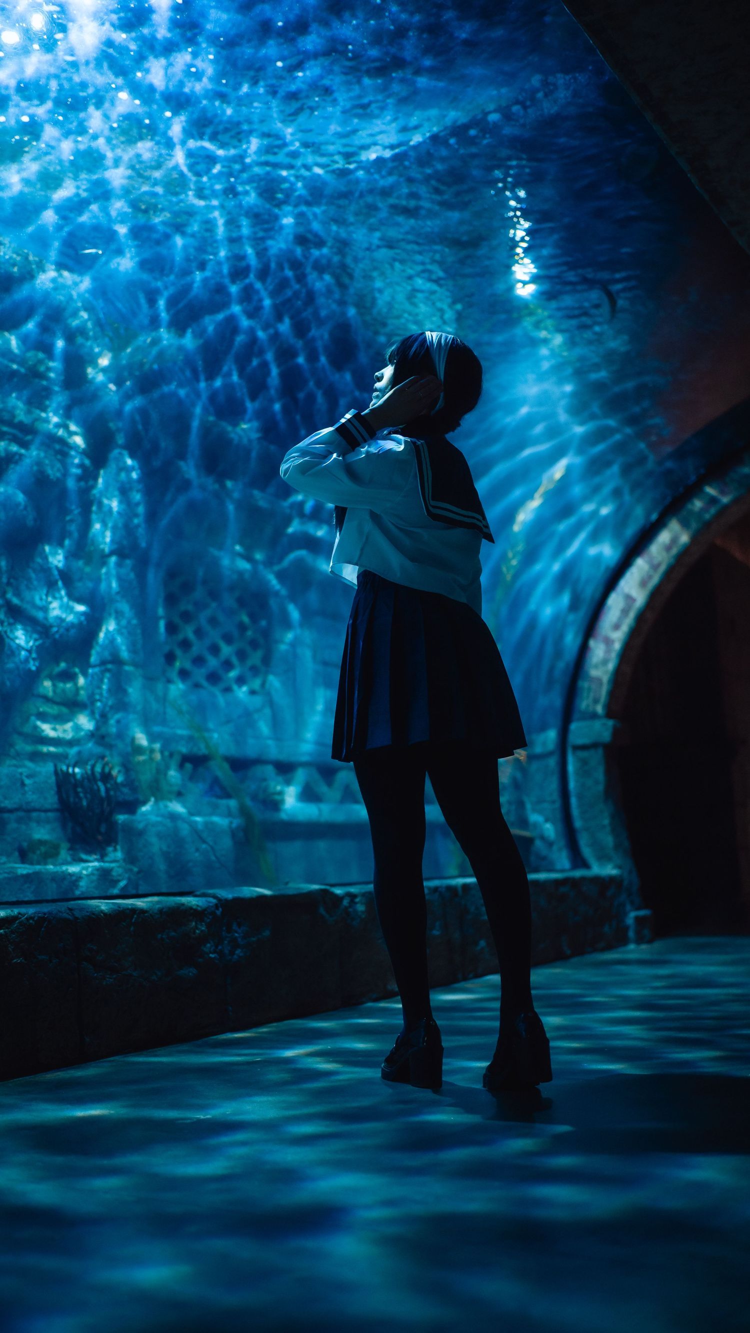 A girl in a Japanese school uniform stands in a dimly lit aquarium tunnel, bathed in blue light from the tank above.