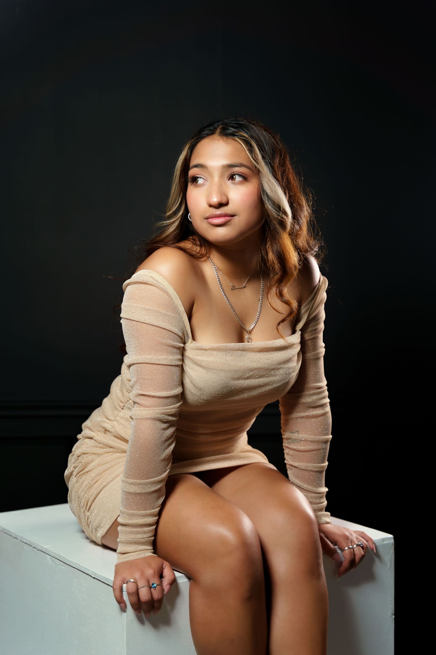 Young woman in beige off-shoulder dress sitting on white cube against dark background.