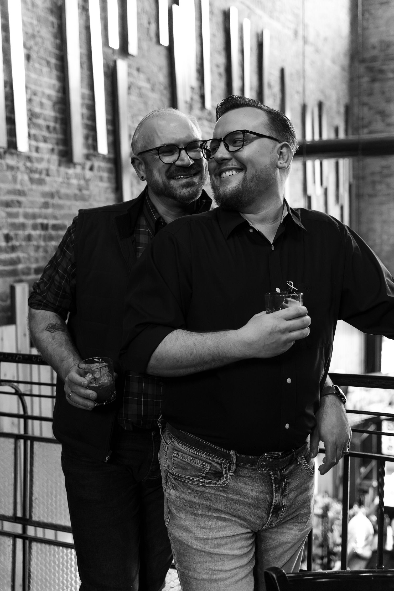 Two smiling men wearing glasses embrace and hold drinks in a black and white photo at an industrial venue.