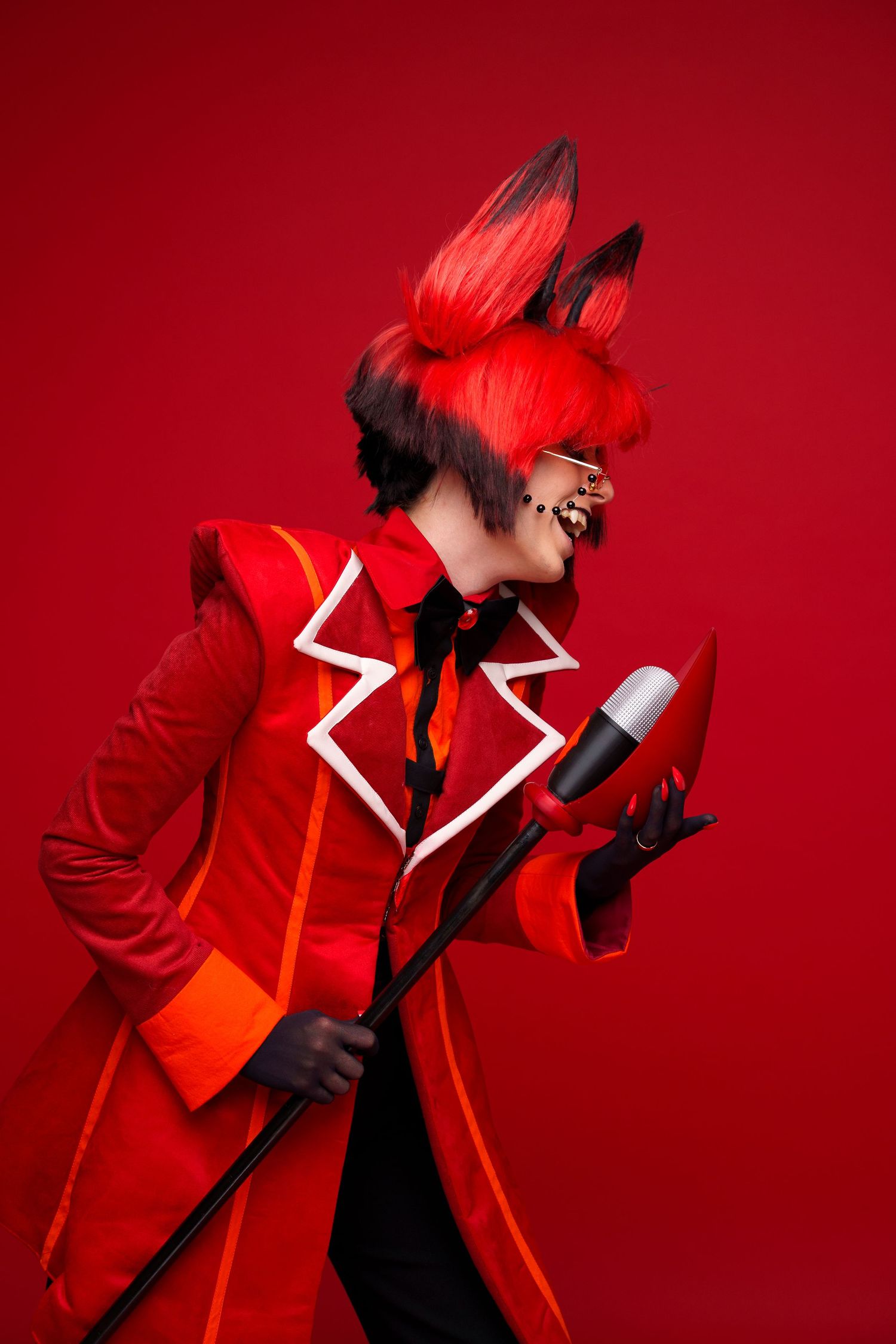 Person in red devil costume with dramatic makeup, bunny ears, and microphone performing on red background.