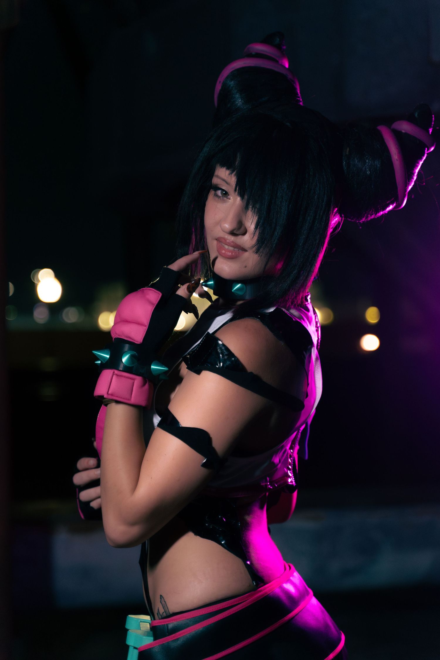 Woman in pink and black anime cosplay costume with cat ears poses dramatically under colorful stage lighting.