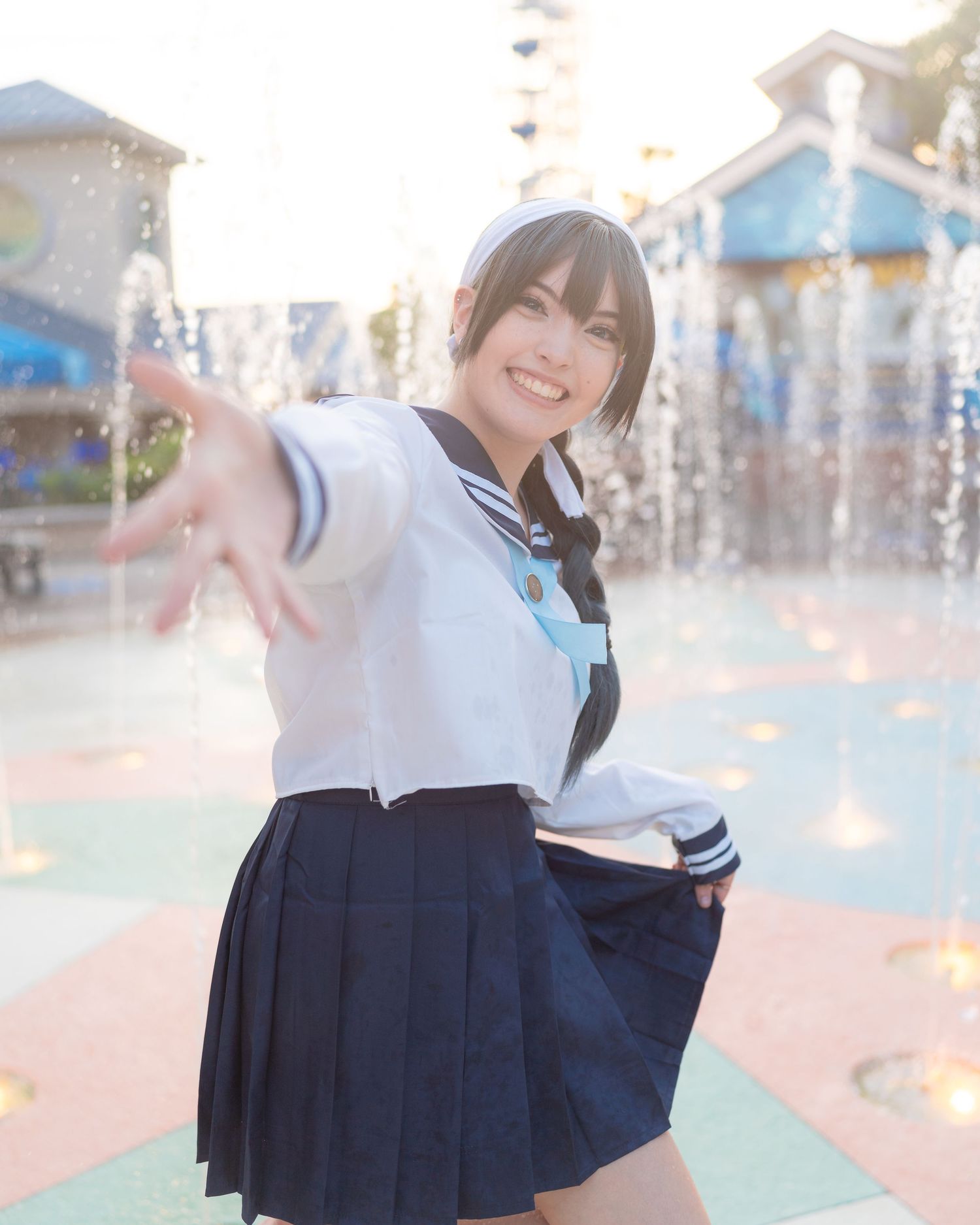 Young woman in Japanese sailor school uniform smiling and reaching toward camera near fountain outdoors.