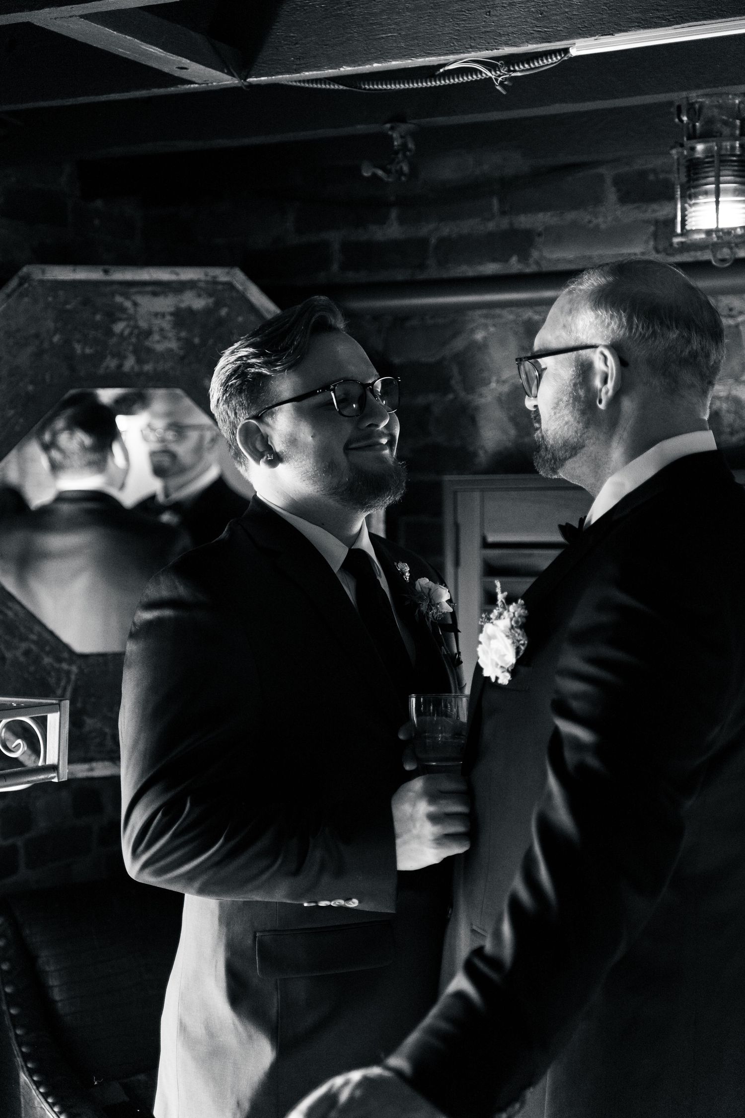 Two grooms in tuxedos share a joyful moment at their wedding reception in a dimly lit venue.