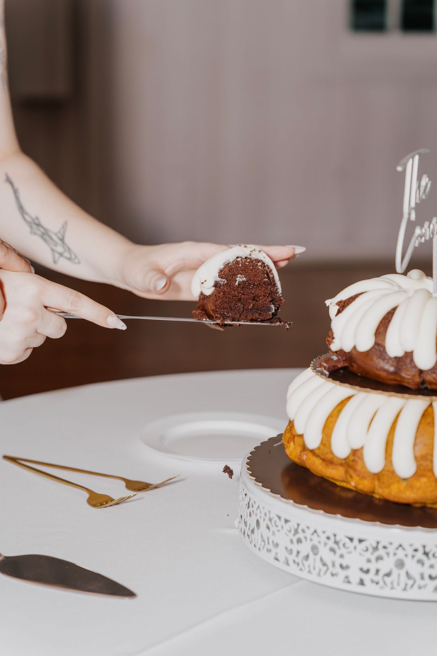A tattooed hand lifts a slice of chocolate bundt wedding cake with white frosting from a two-tier cake on a decorative stand.