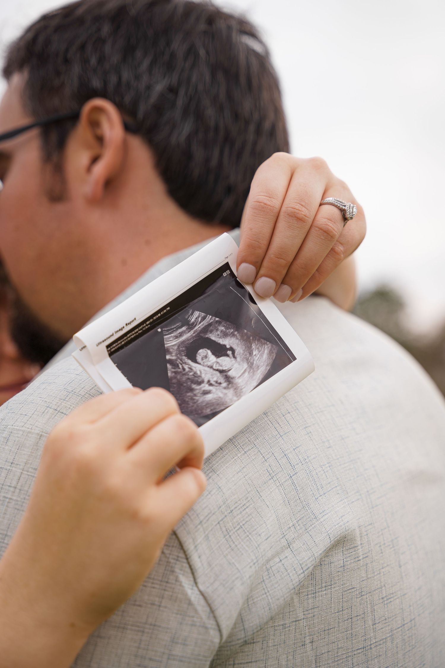 Couple embracing while woman holds ultrasound photo over man's shoulder, announcing pregnancy with engagement ring visible.