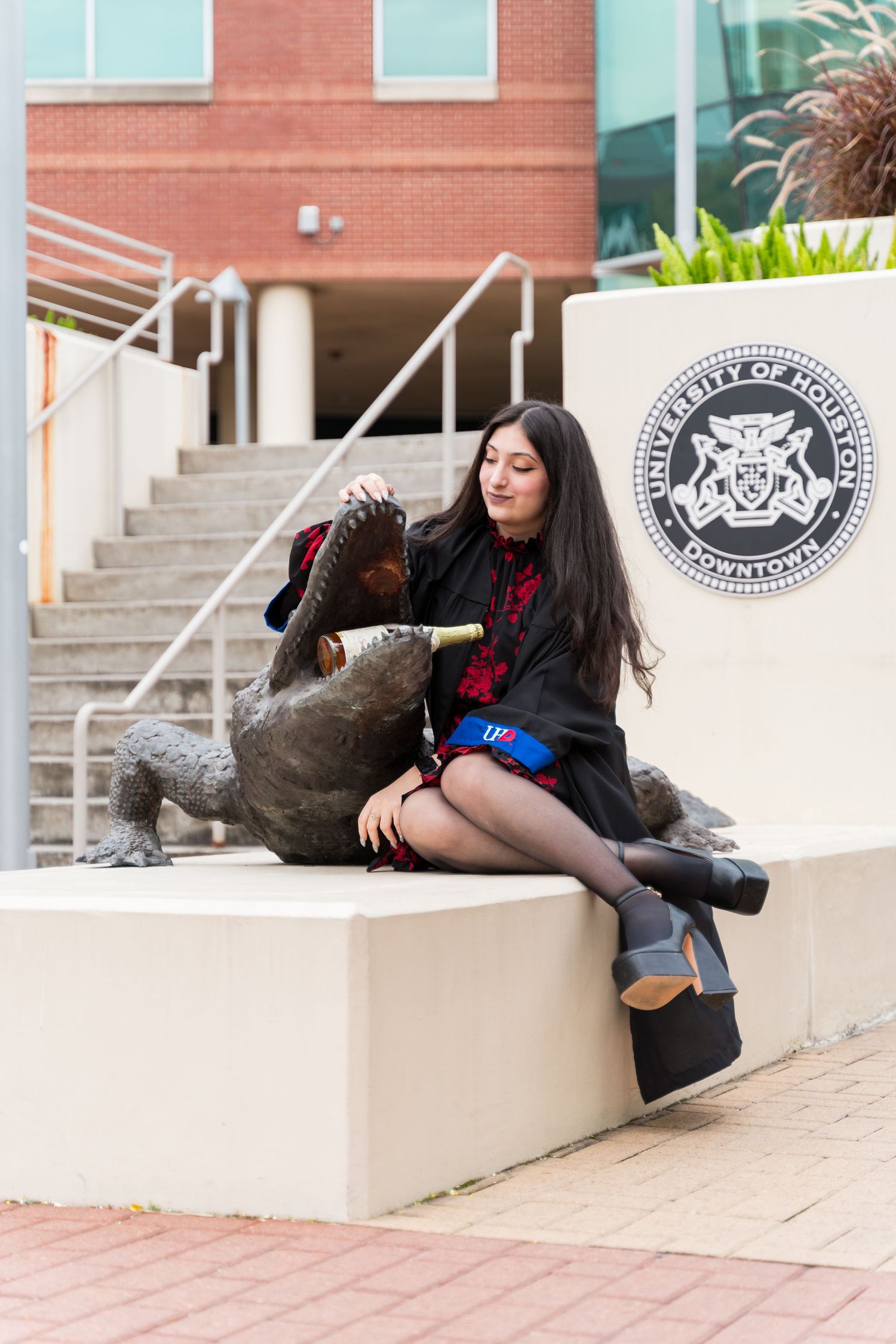 Graduate in black gown poses with gator statue at University of Houston Downtown campus.