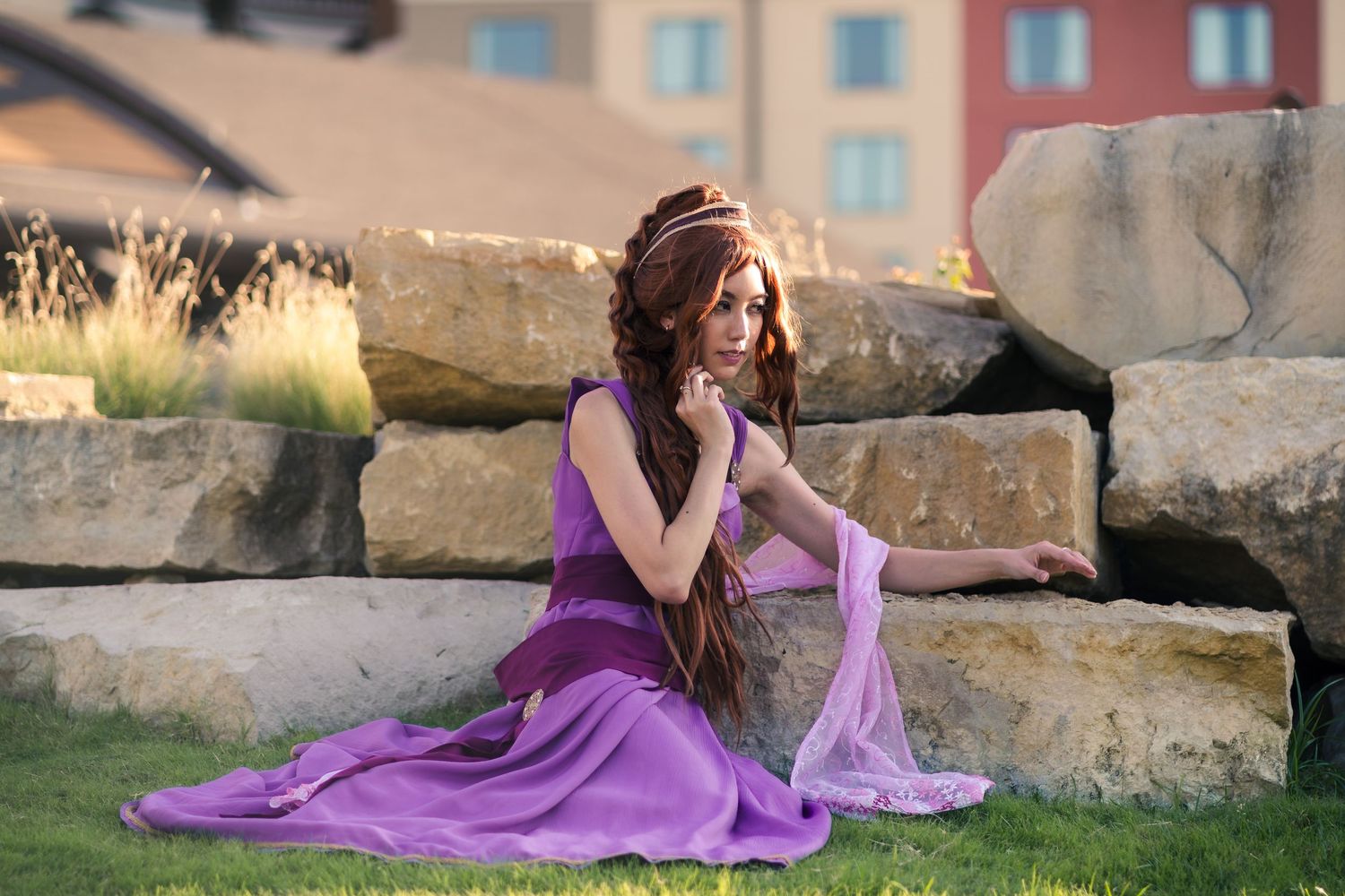 A woman in a flowing purple gown with long red hair sits gracefully on stone steps outdoors.