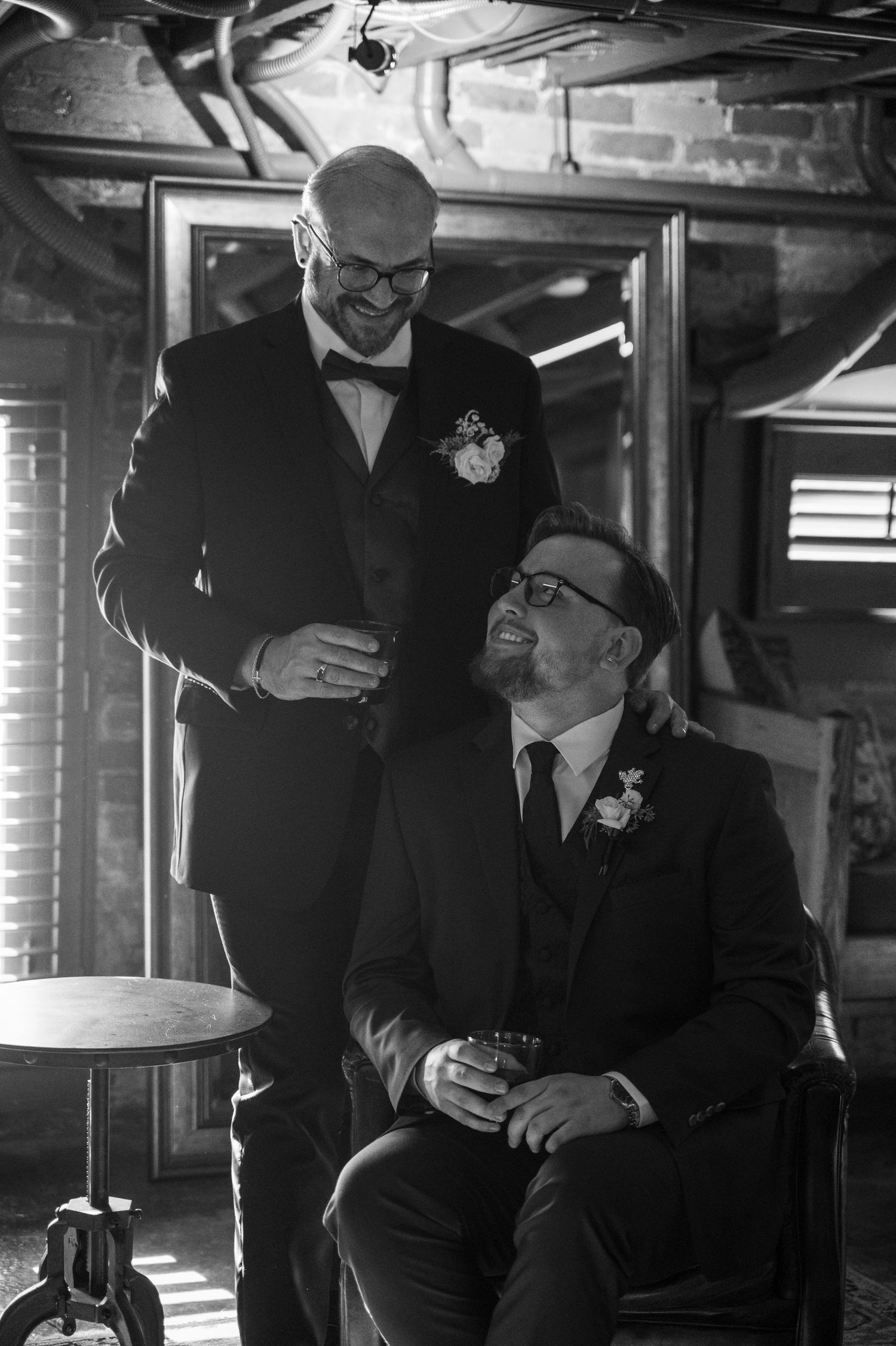 Two people in formal attire share intimate moments in a dimly lit space with brick walls.