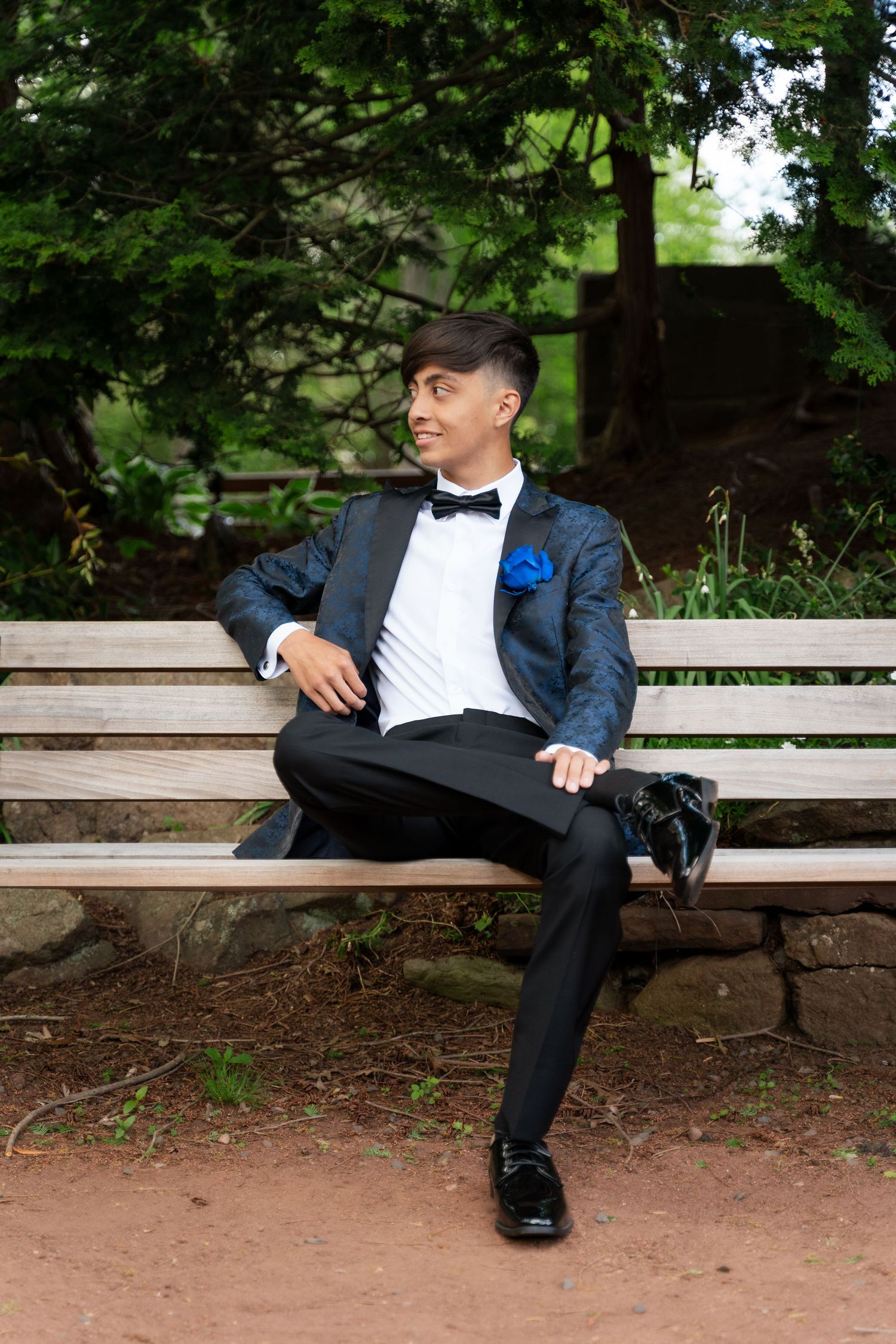 Young man in tuxedo with blue boutonniere sits on wooden bench in garden setting.