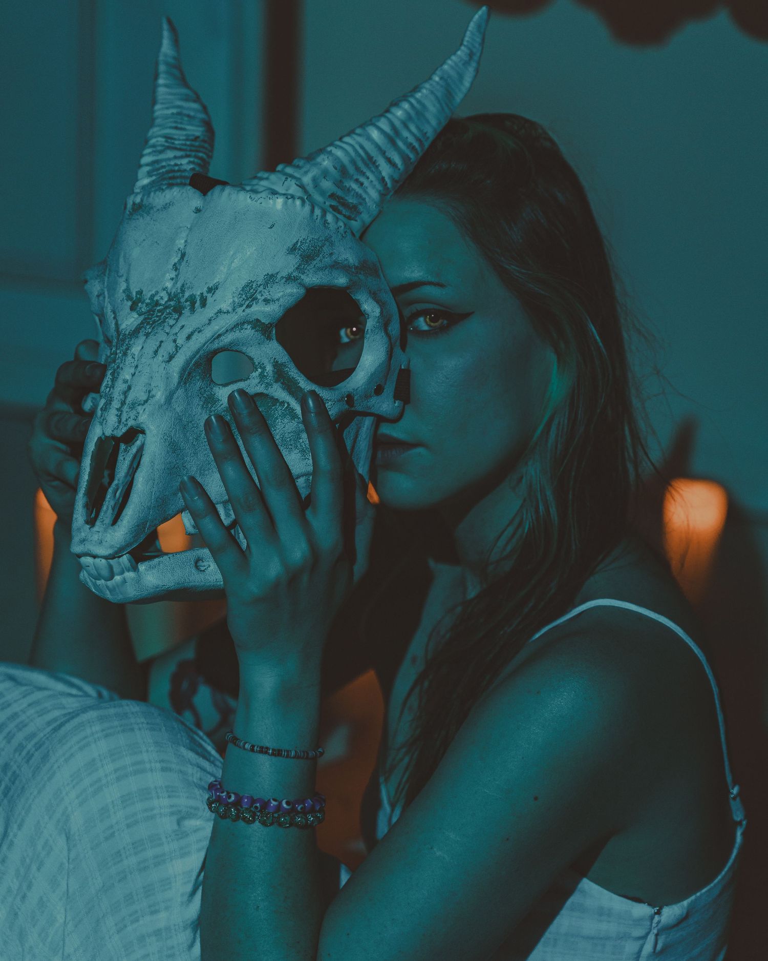 Woman holding a horned animal skull up to her face in moody teal-toned atmospheric lighting.