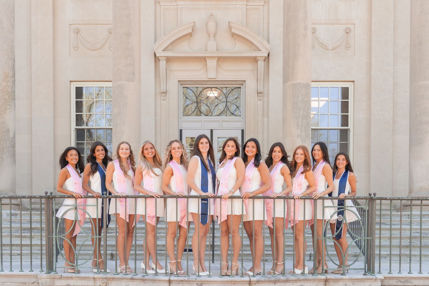 Group of young women in pink and white outfits posing on a balcony in front of a classic stone building.