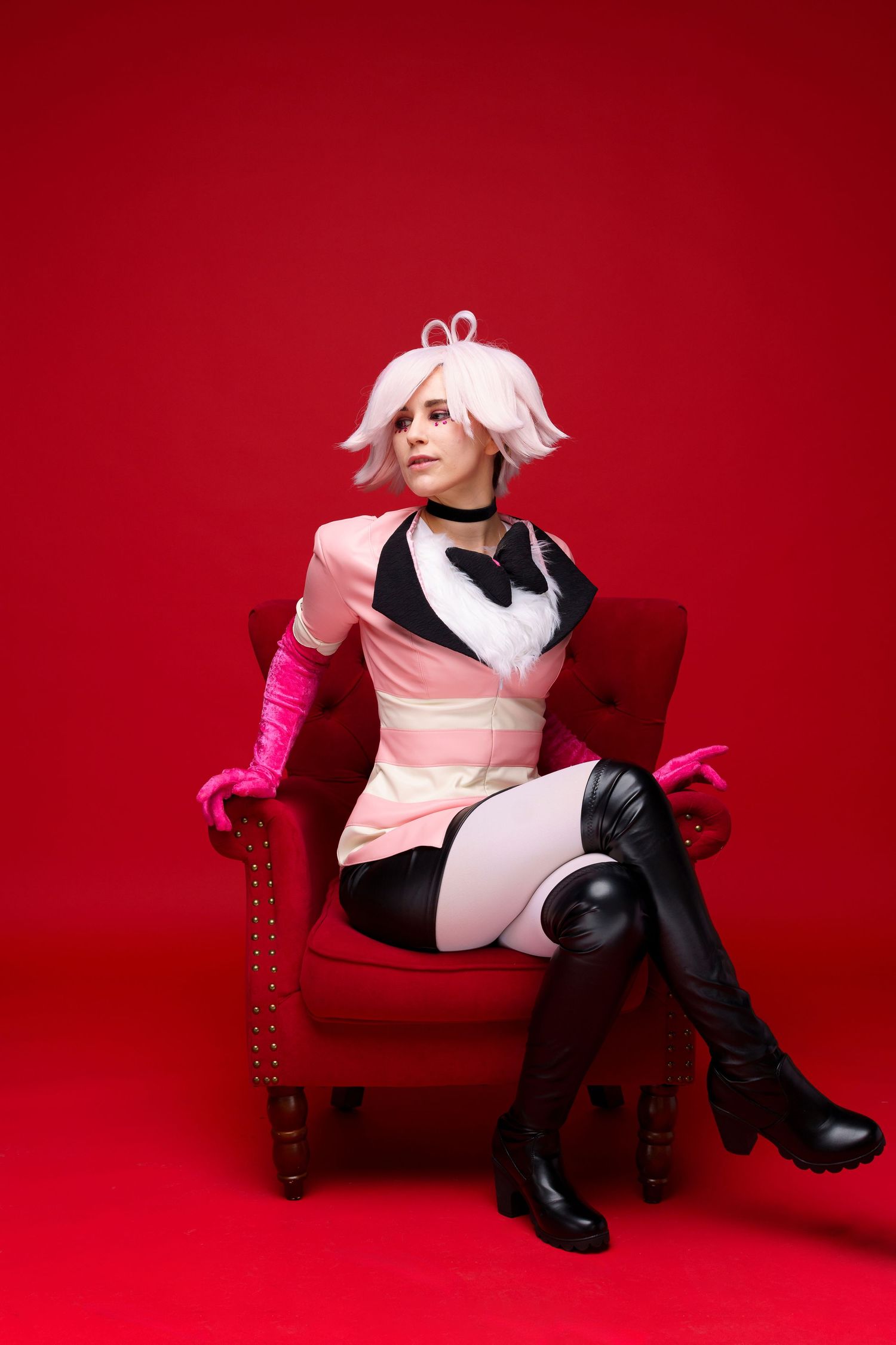 Woman in pink anime cosplay costume with white wig sits on red velvet chair against red background.