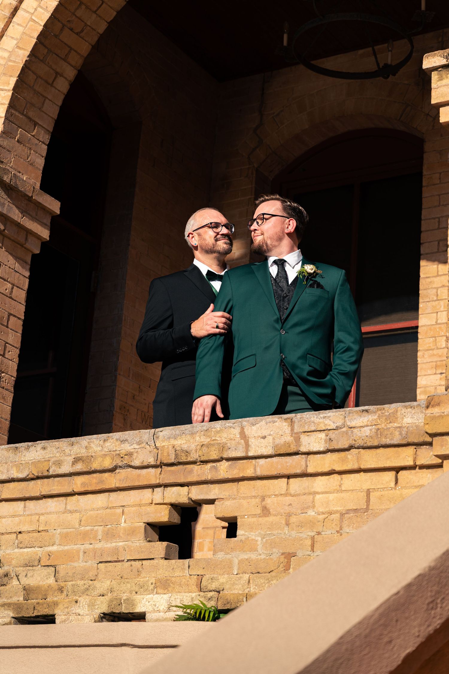 Two grooms in green and black suits stand on a brick balcony, sharing a joyful moment at their wedding venue.