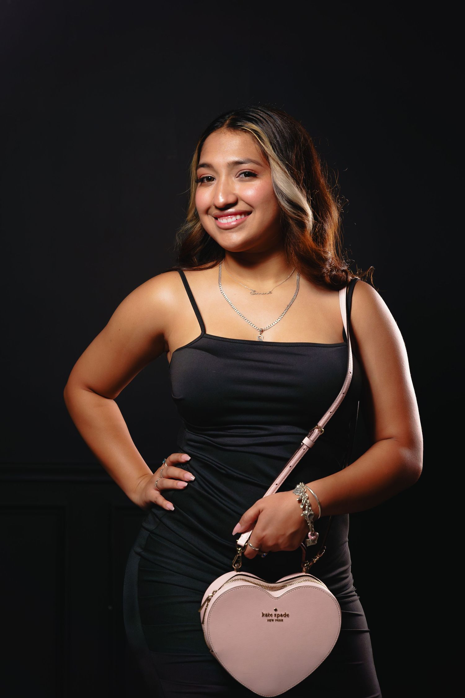 Young woman in black dress holding pink Kate Spade heart-shaped bag, smiling against dark background.