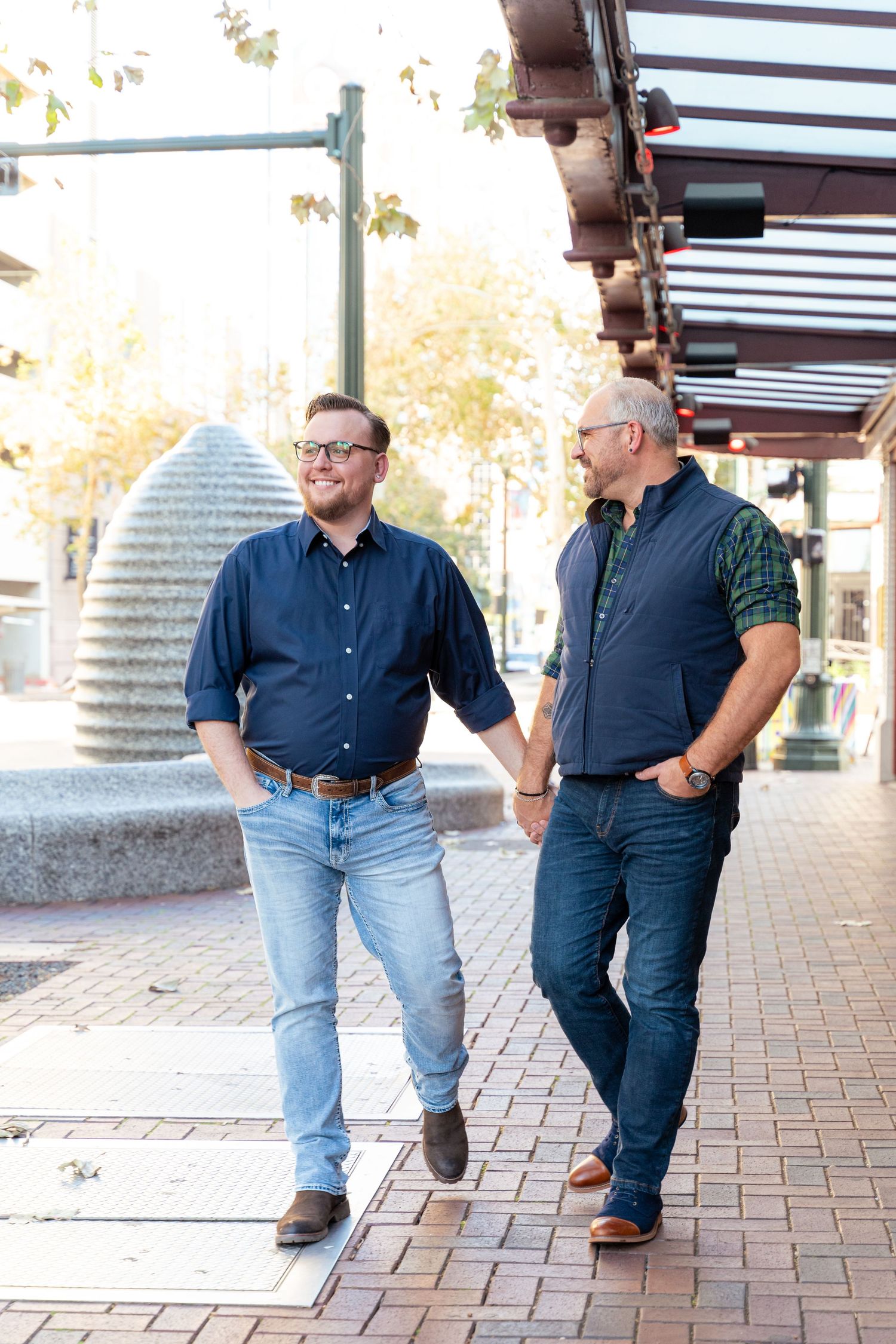 Two men holding hands and walking together on a brick sidewalk in an urban setting on a sunny day.