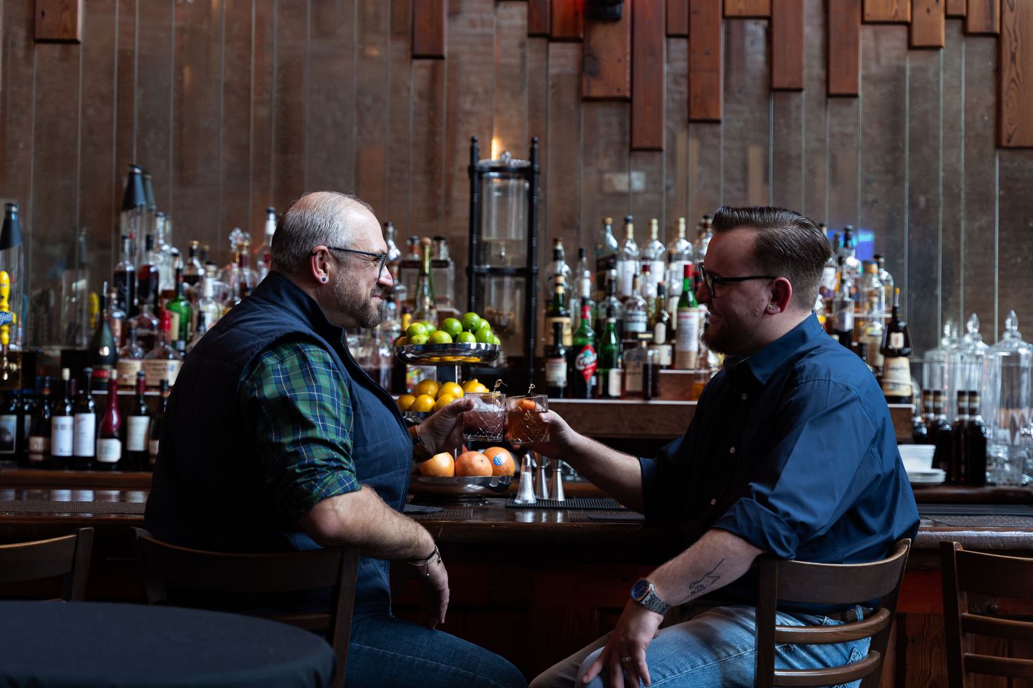 Dark moody bar scene with bottles displayed on wooden shelving and ambient lighting.