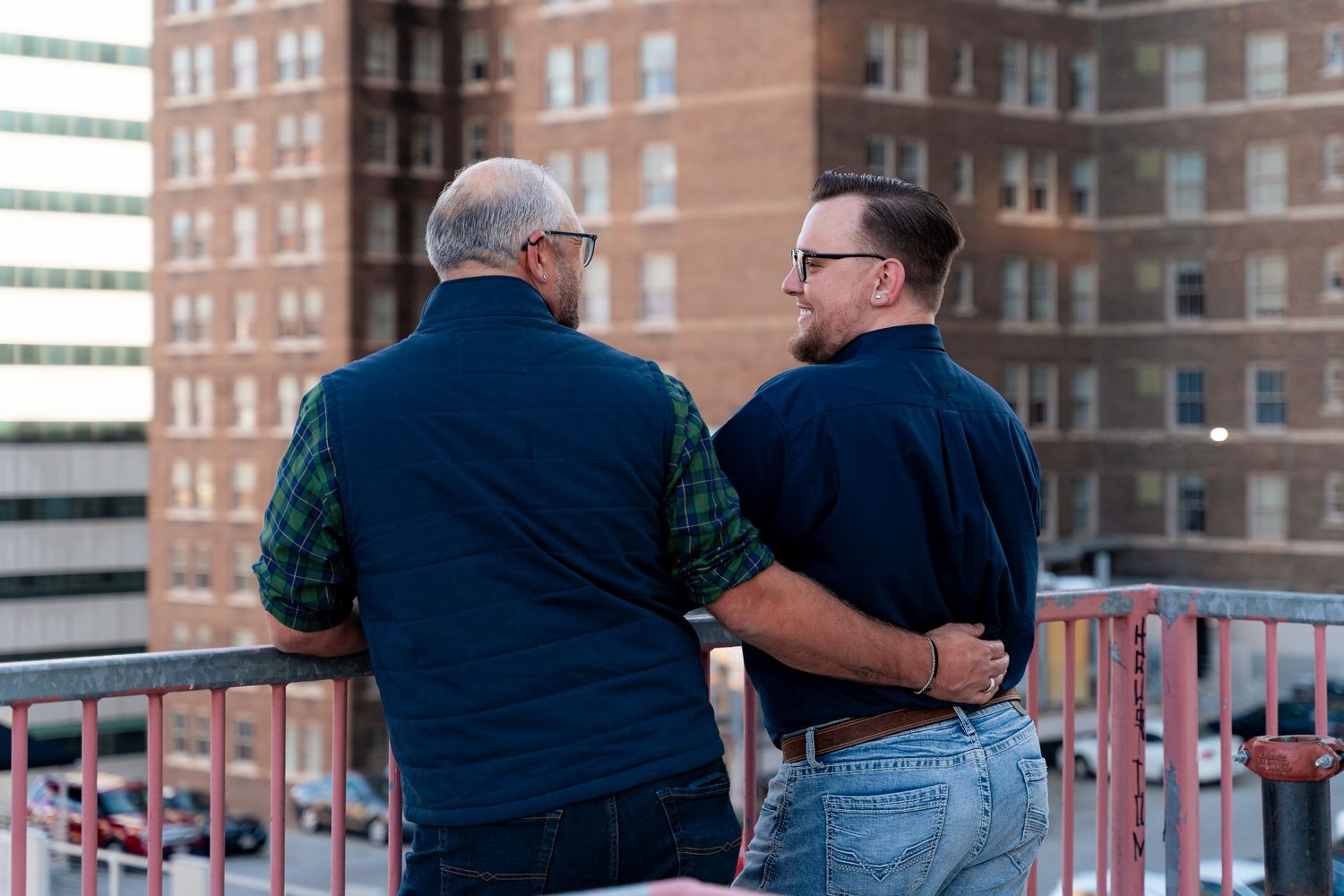 Two men standing on a rooftop balcony overlooking city buildings, viewed from behind.