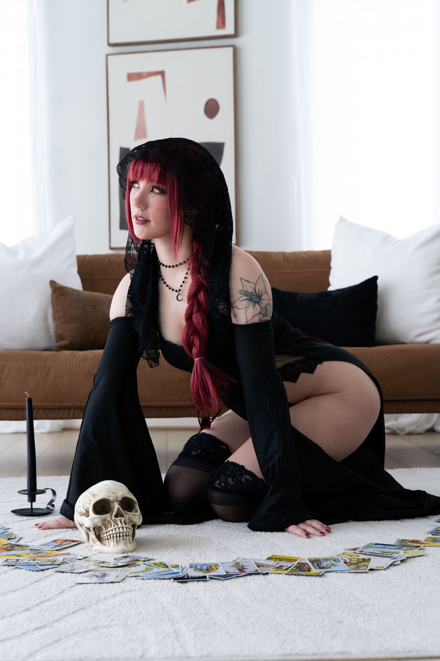 Woman in black gothic costume with red hair sits on floor surrounded by tarot cards and skull prop.