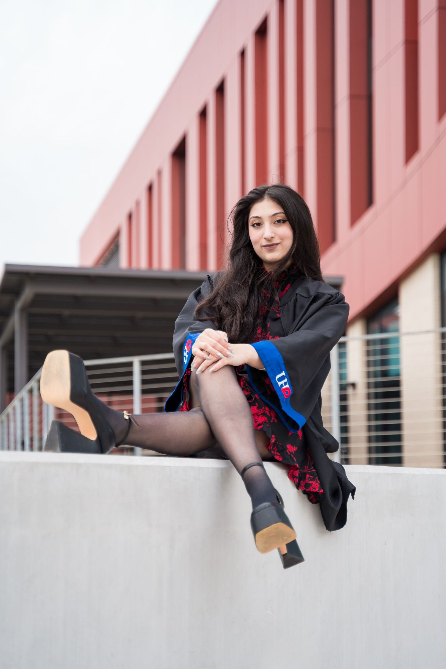 A young woman in graduation gown and blue sash poses confidently on a concrete wall outside a modern red building.