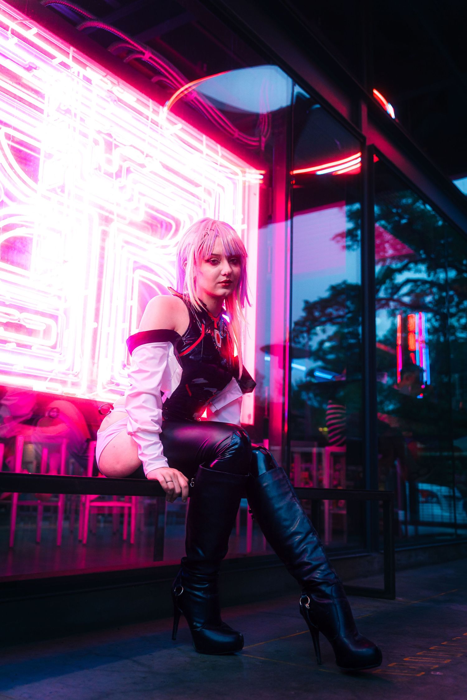 Woman in cyberpunk cosplay with white hair, black leather outfit, and thigh-high boots poses by neon pink lights.