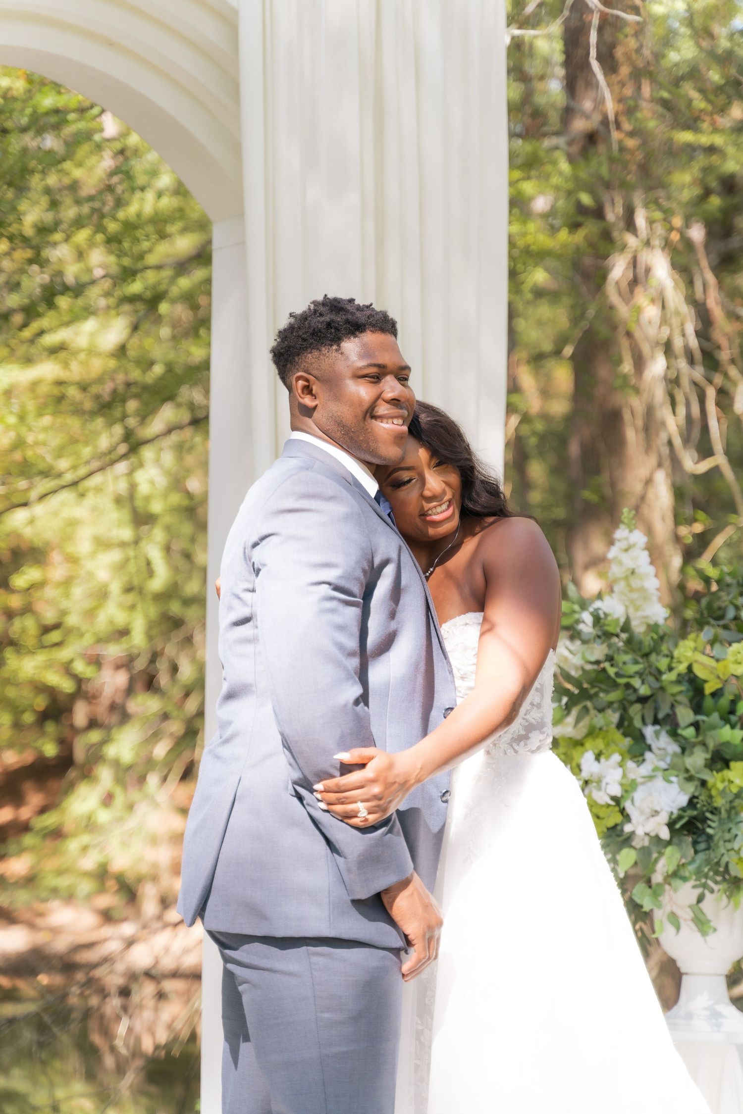 A joyful bride and groom embrace outdoors near white columns, surrounded by lush greenery on their wedding day.