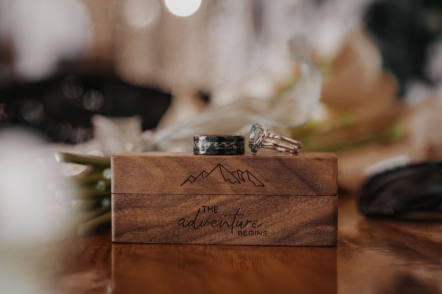 Wedding rings resting on a wooden box engraved with mountains and the phrase The Adventure Begins.