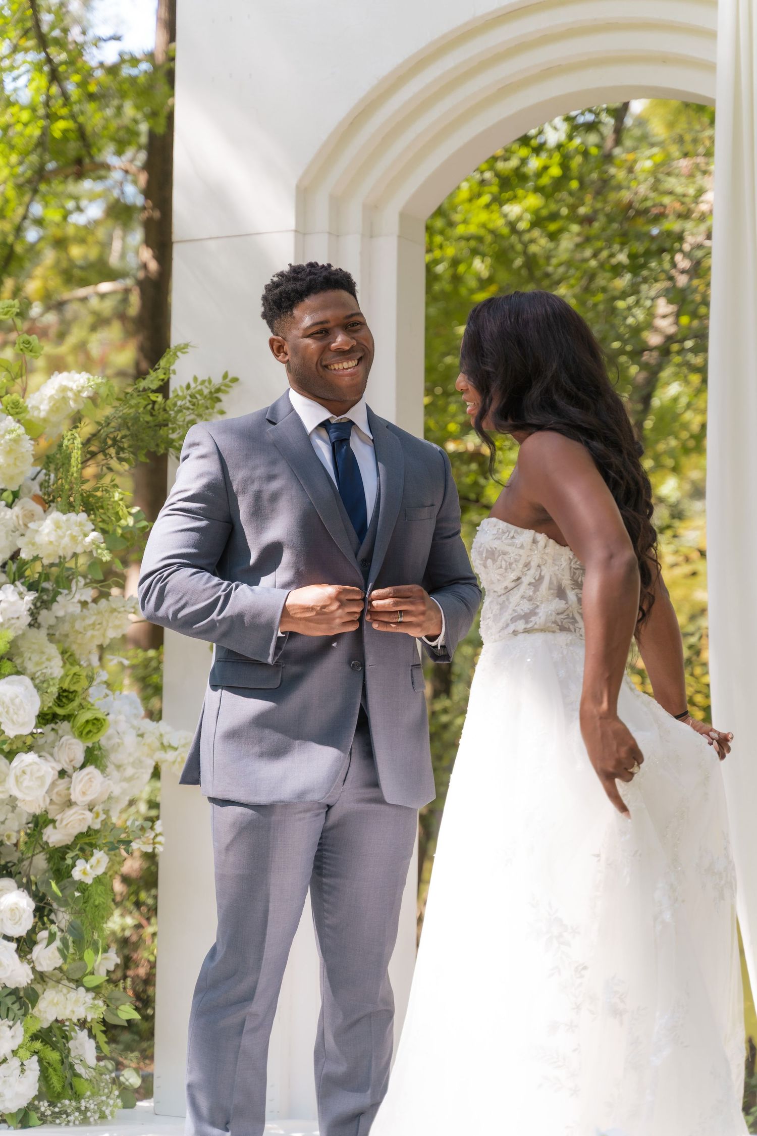 A couple shares a tender moment during their wedding ceremony under an archway decorated with white flowers.