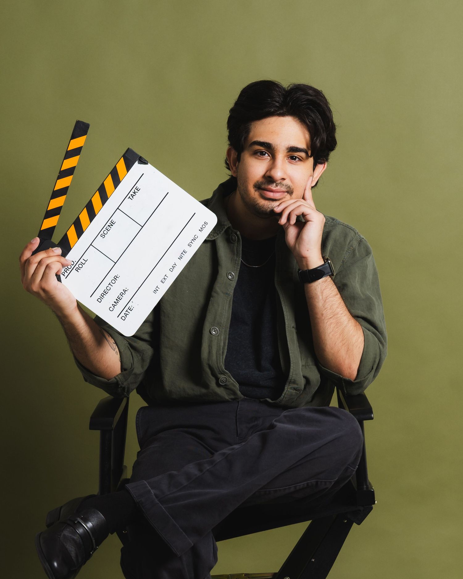 Young filmmaker holding a clapperboard on a director's chair against an olive green background.