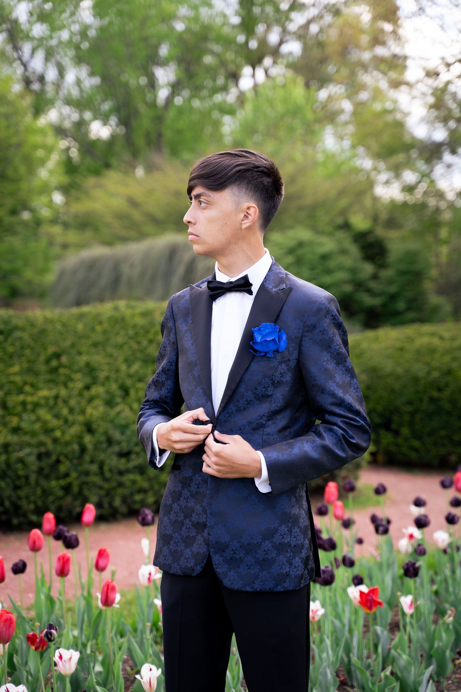 Young man in patterned navy tuxedo with blue boutonniere and bow tie poses in garden with colorful tulips.