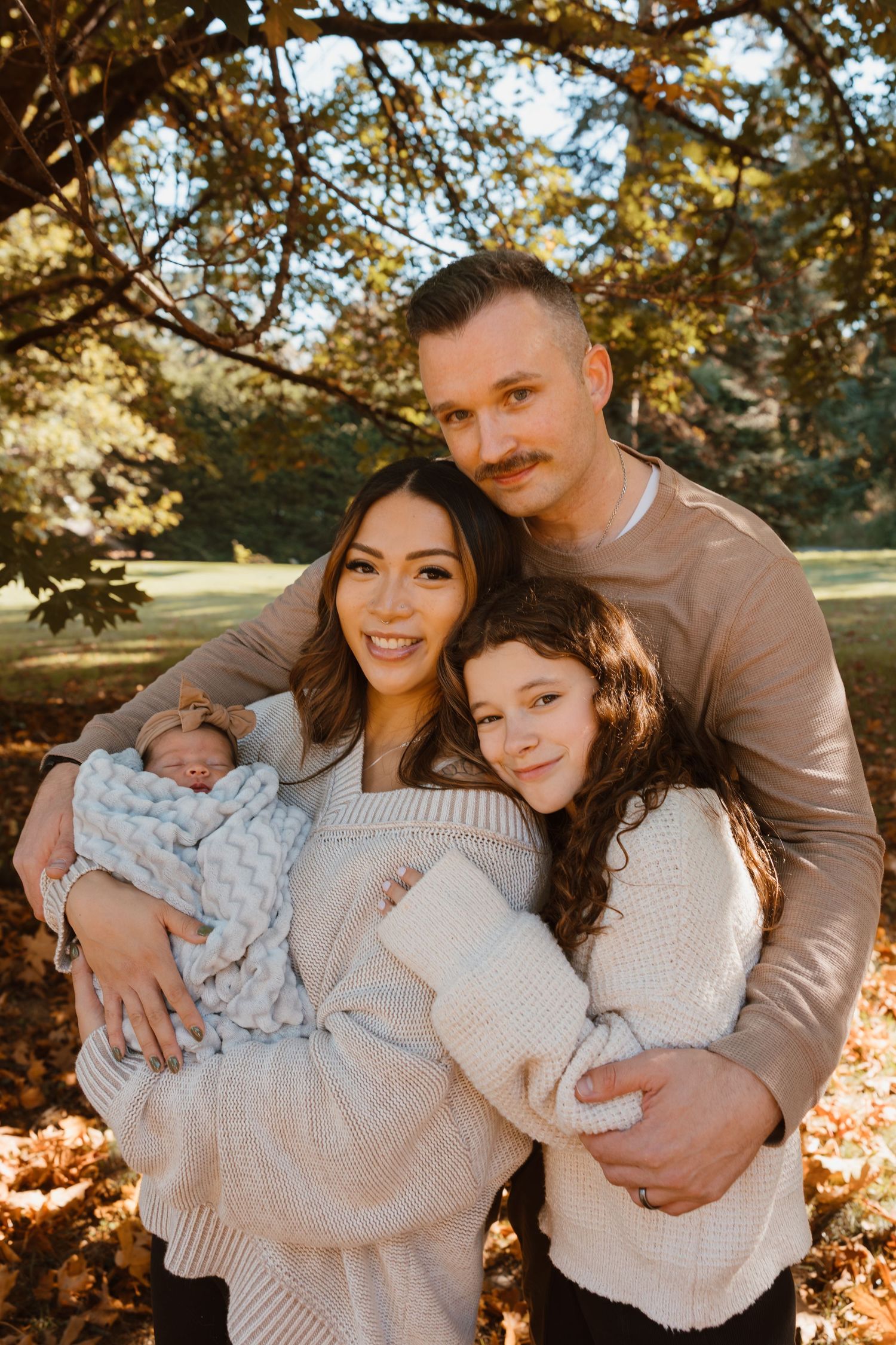 Family cuddles together on autumn day with newborn baby wrapped in blue blanket under golden sunlit trees.