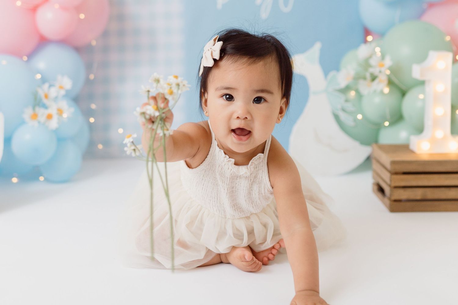 A baby in white dress crawls during a birthday photoshoot with pink and blue balloons.