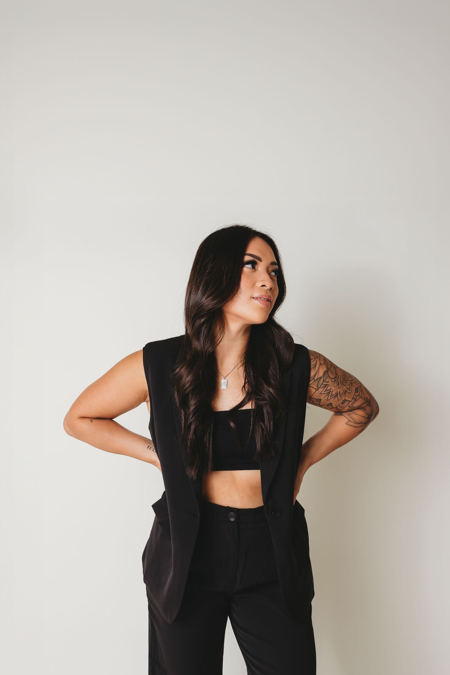 Woman in black crop top and vest poses with hand on hip against white wall.