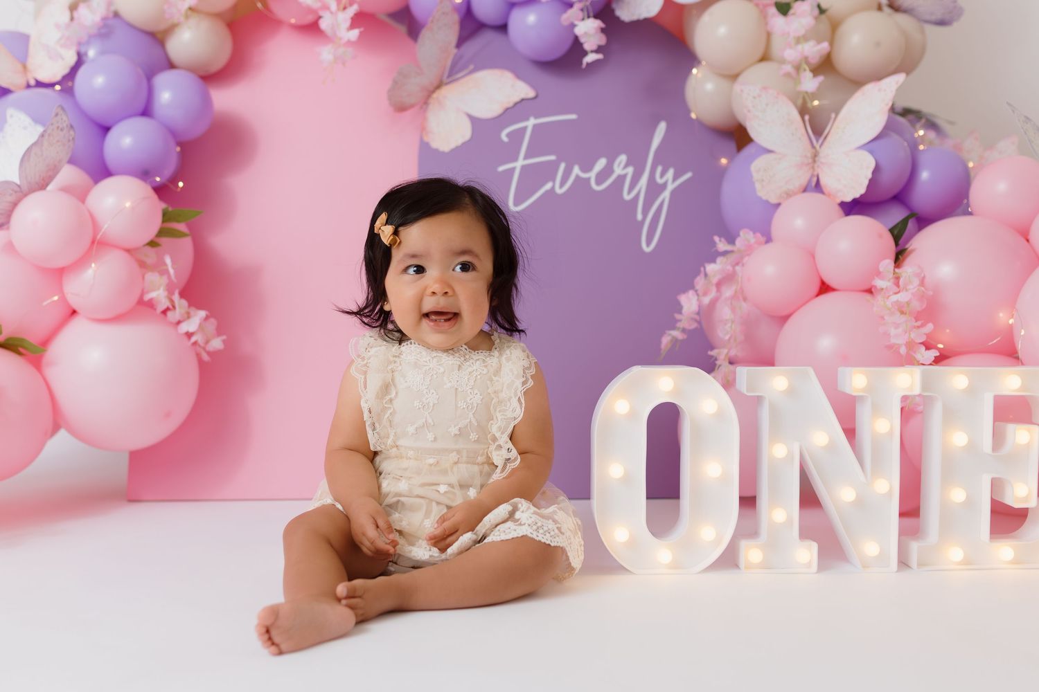 A baby sits by illuminated ONE sign with pink and purple balloon arch decorations.