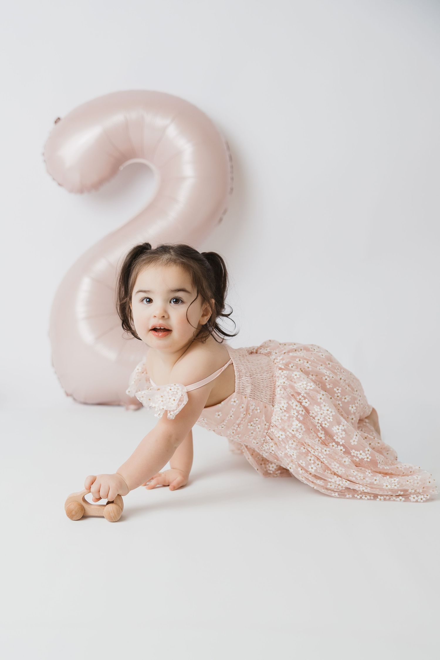 Toddler girl in pink lace dress crawling near a number 2 balloon, celebrating her 2nd birthday.