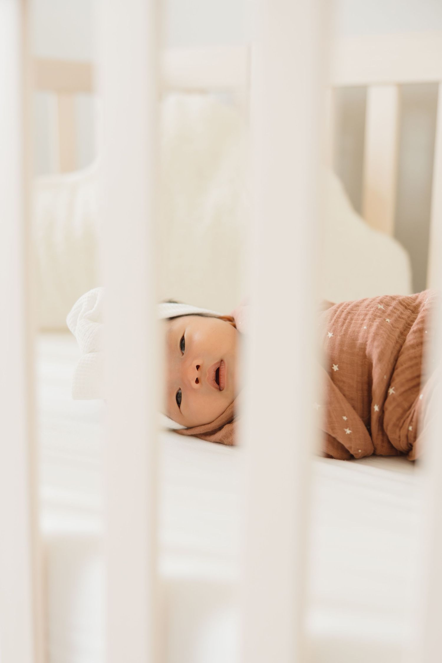 Close-up view of a sleeping baby through white crib bars in natural light.