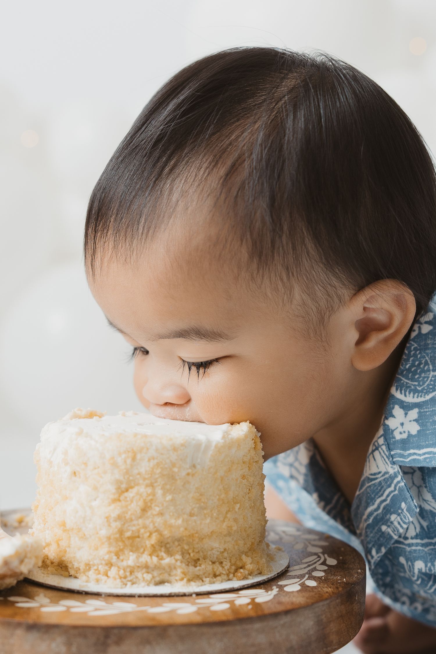 Cute Asian toddler leaning in to take a bite of a white frosted birthday cake on a wooden stand.