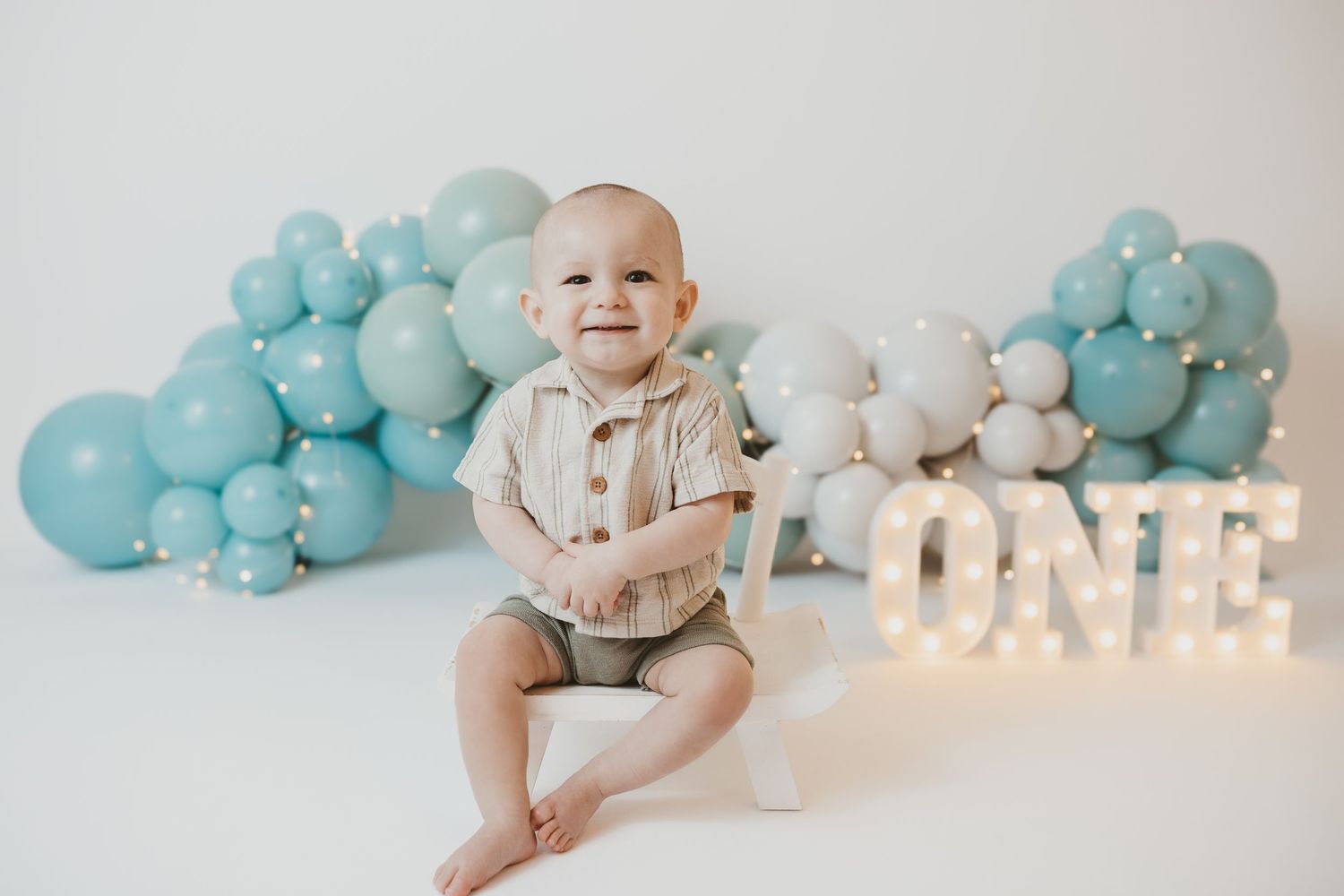 A baby sits beside lit ONE sign with blue and white balloon decorations.