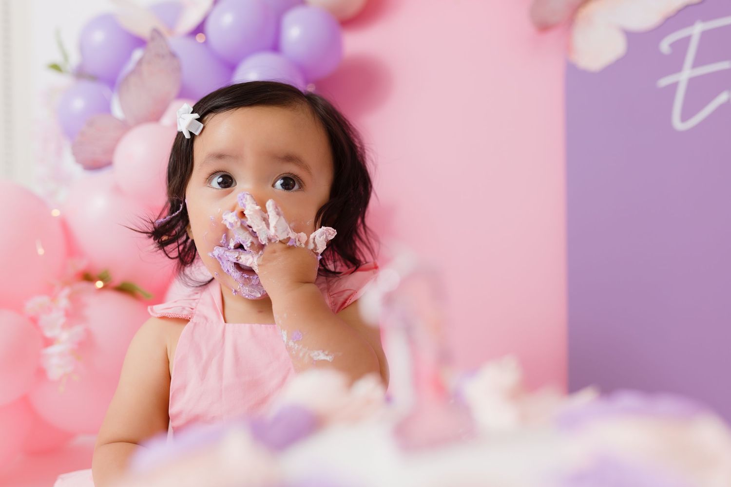 A baby in pink dress plays with confetti during a birthday photoshoot with purple and pink balloons.