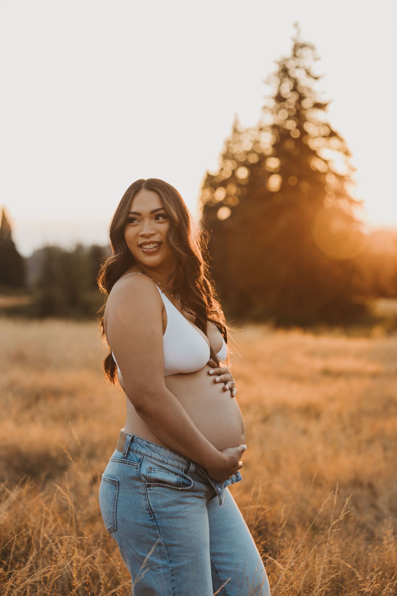 Expectant mother in denim and white top poses in golden sunlit field at sunset.