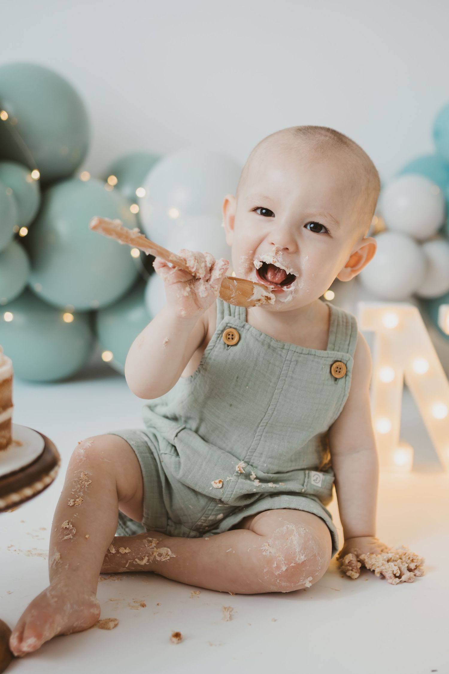 A baby in mint overalls messily eats cake during first birthday photoshoot.
