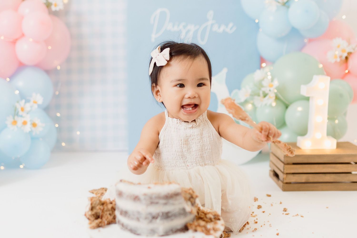 A baby in white dress celebrates with cake and balloons at their first birthday photoshoot.