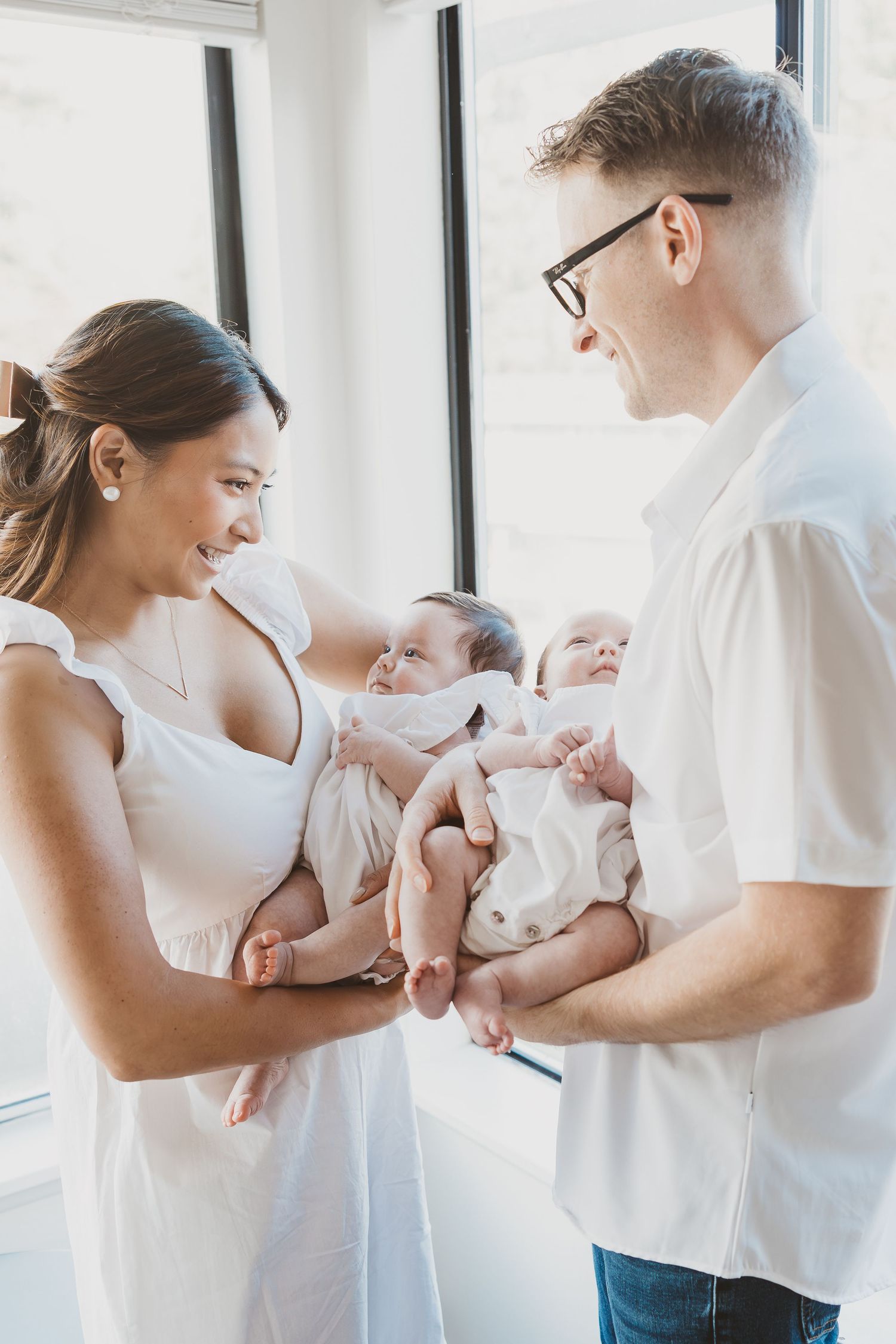 Parents in white clothing holding and playing with their baby by bright windows.