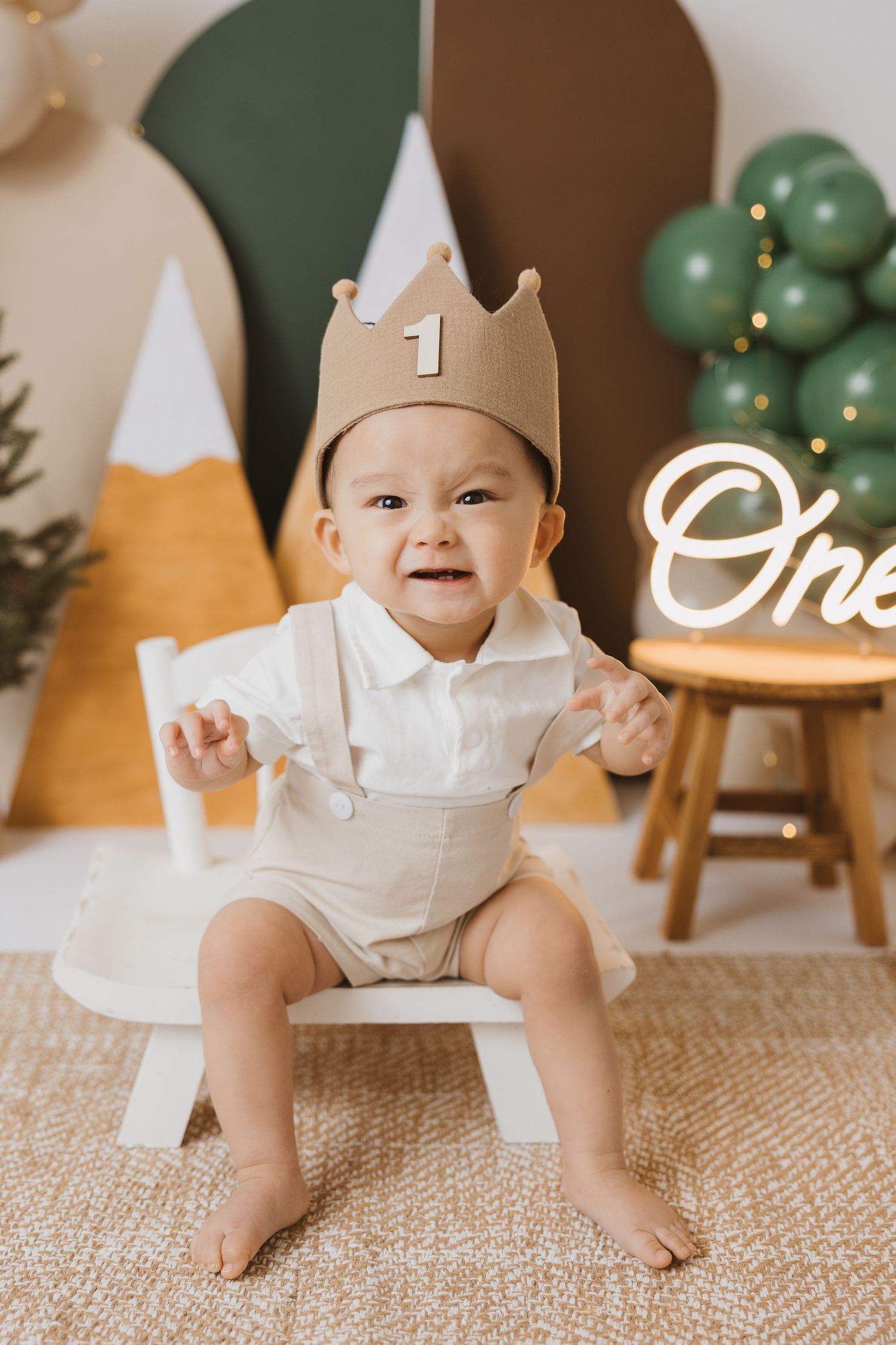 Baby in white outfit crawling on textured rug with birthday decorations and crown.