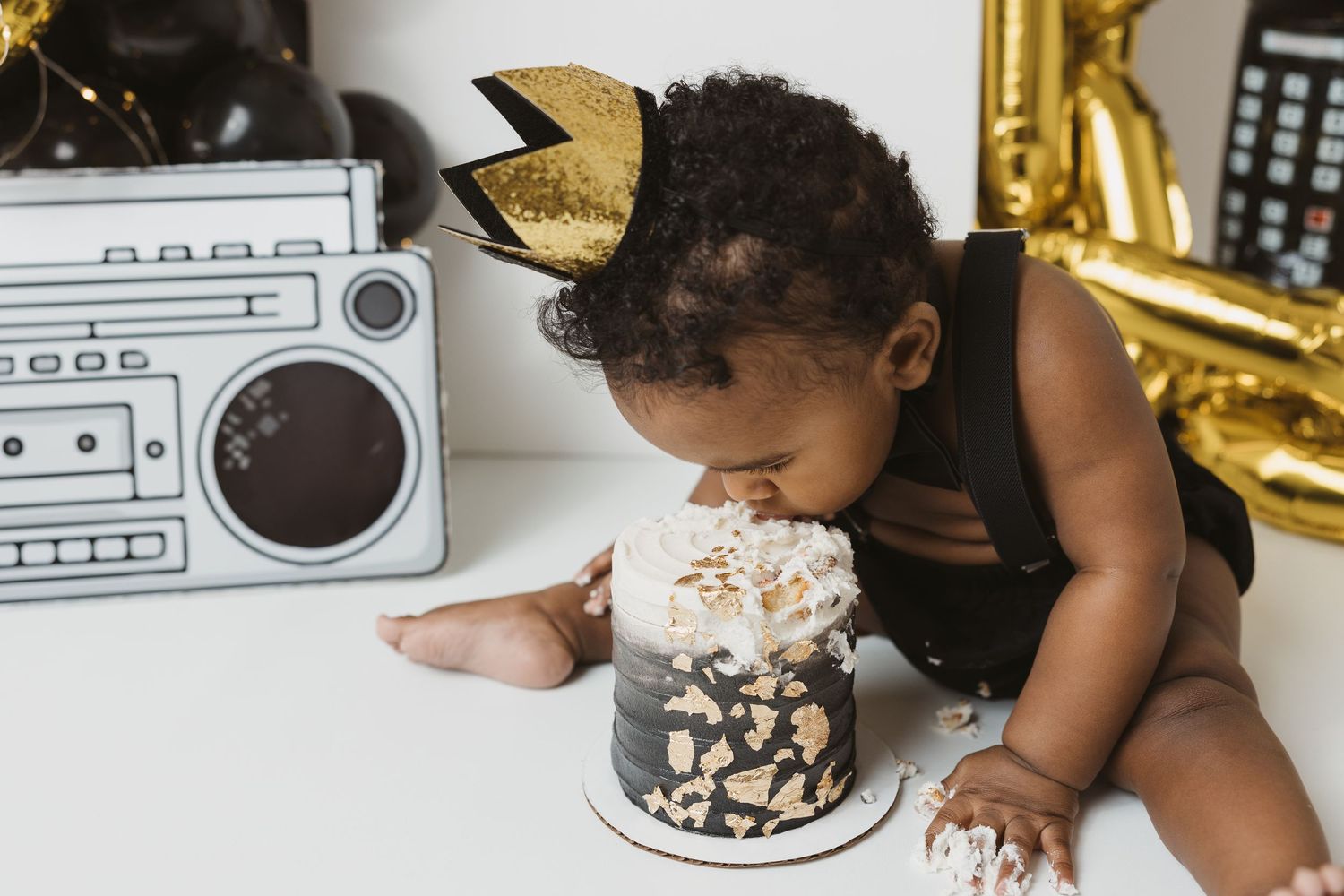 A baby wearing a gold crown headband eating a black and gold smash cake during a first birthday photo shoot.