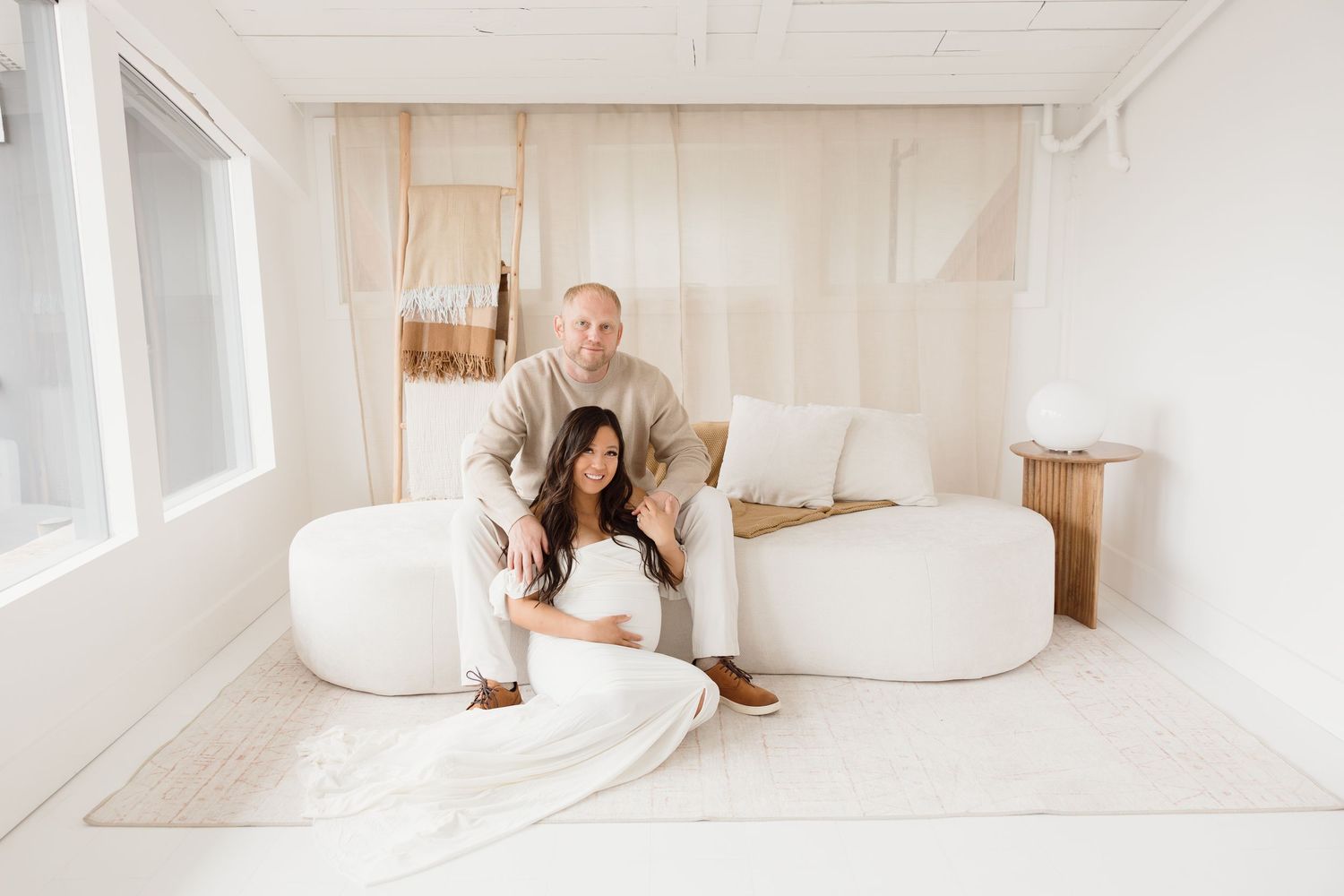 Couple posing in a bright, minimalist white studio, man standing behind seated pregnant woman on a cozy curved sofa.