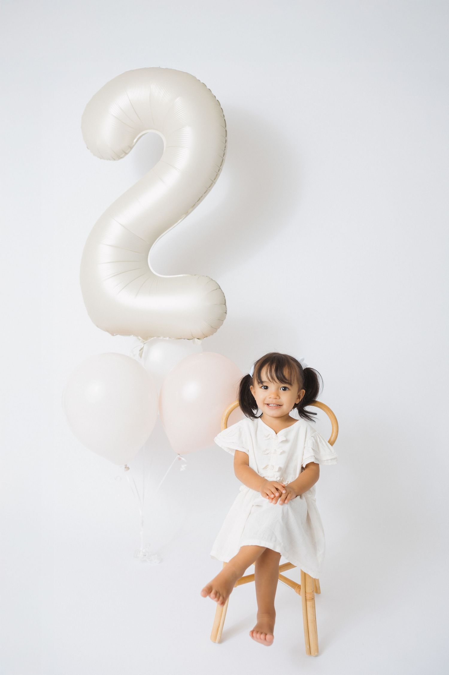 Child in white dress sitting on wooden stool with number 2 balloon.